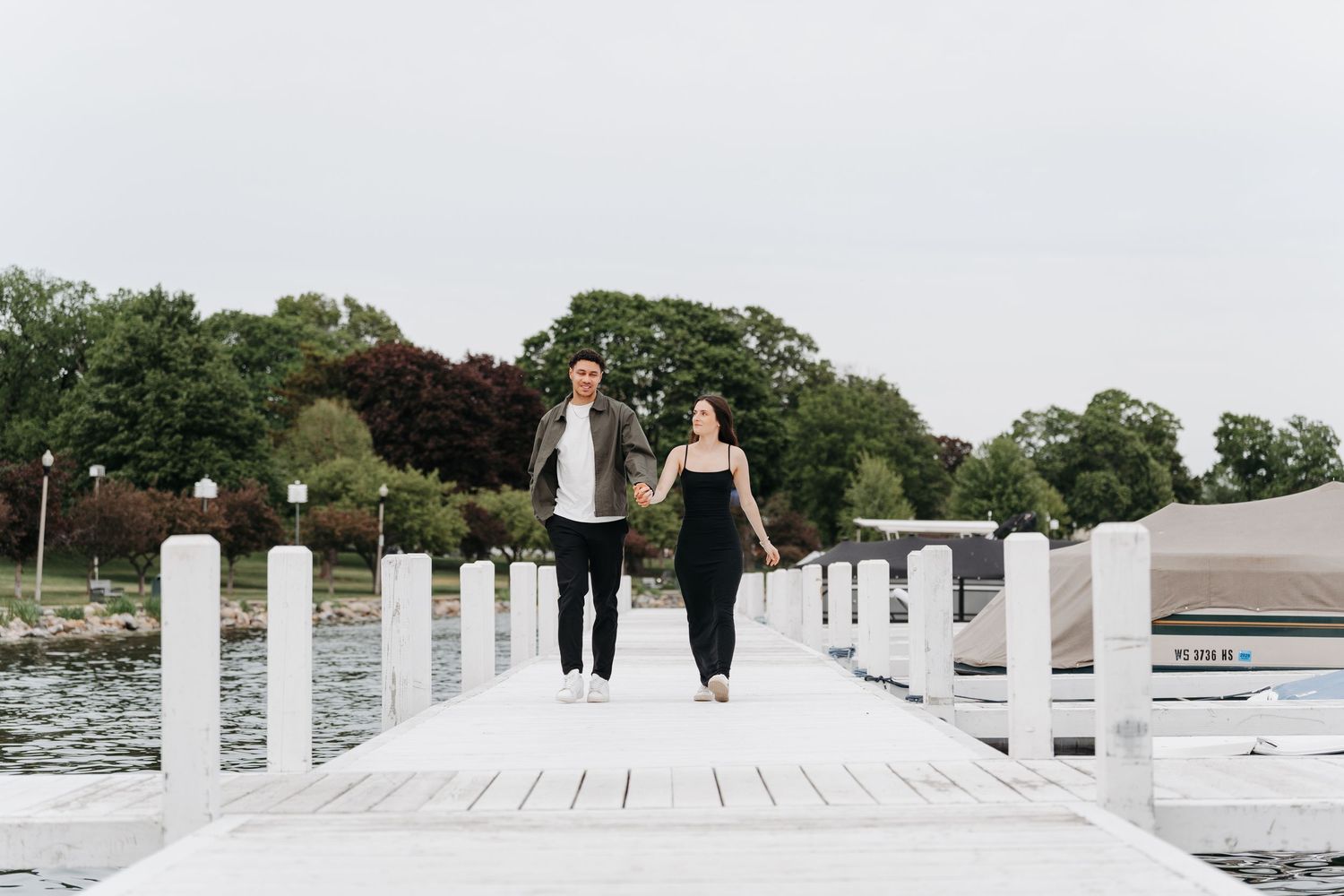 Couple holding hands walking on a white dock by the water, surrounded by lush green trees.