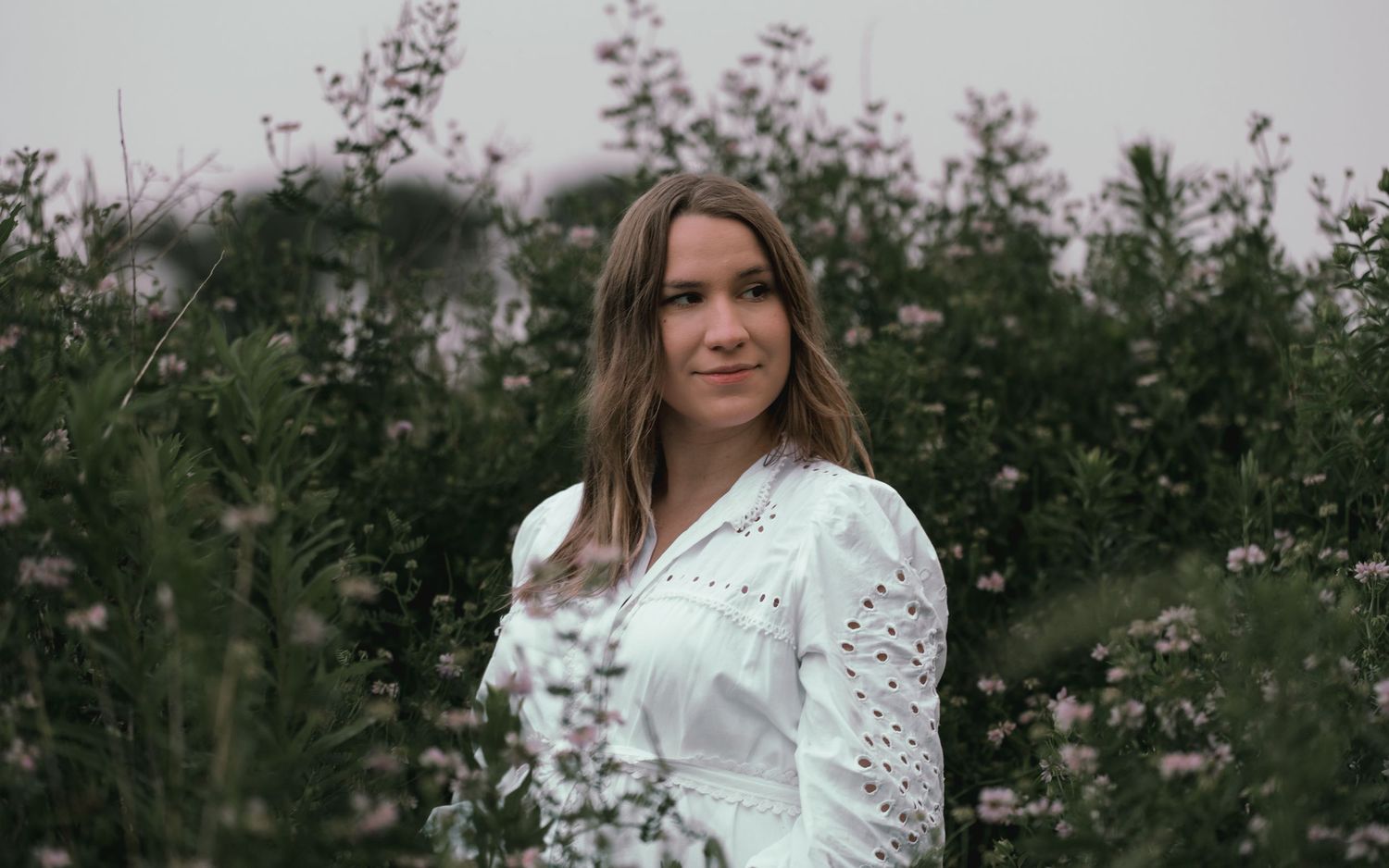 Woman in white eyelet blouse standing among tall pink wildflowers on an overcast day.
