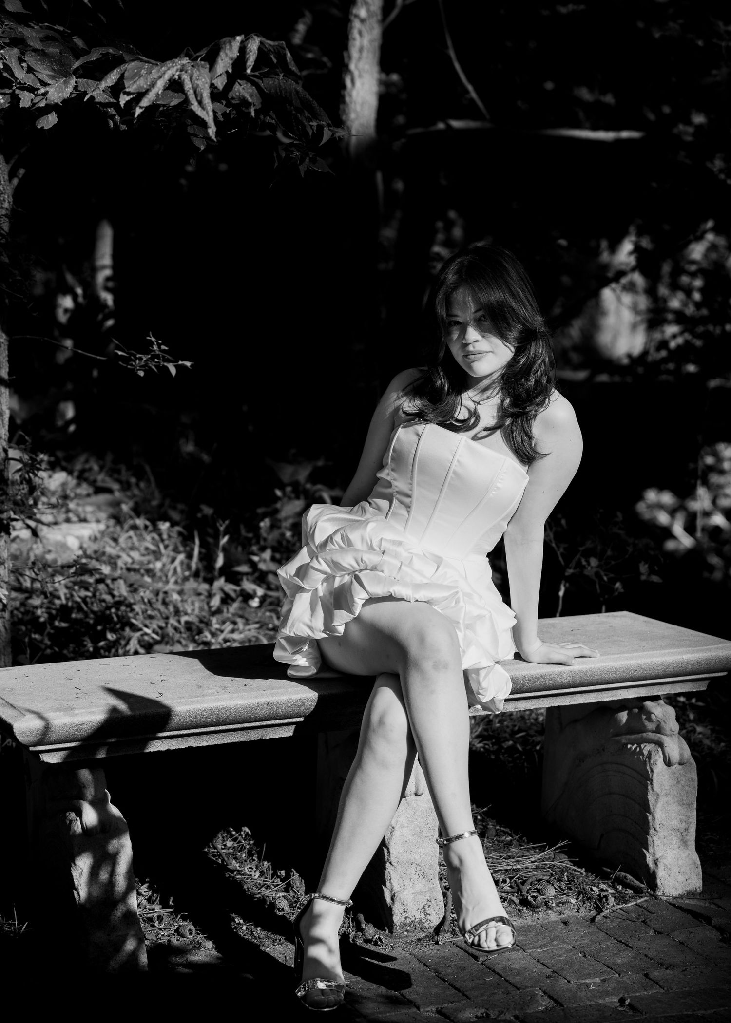 Black and white photo of young woman in white dress sitting on stone bench in garden.
