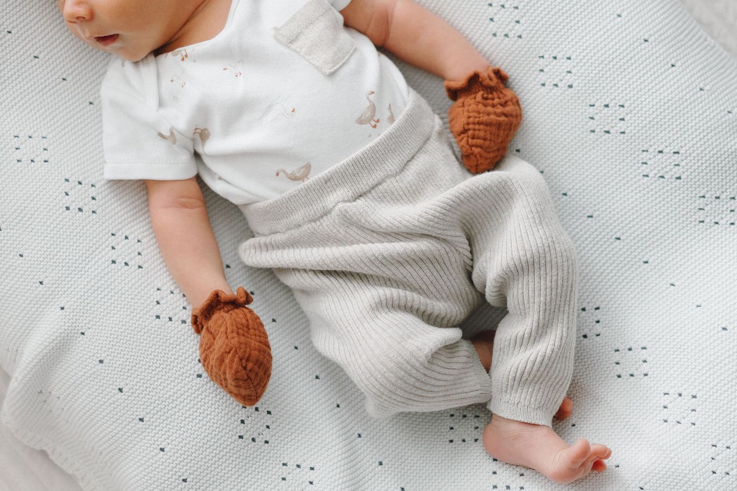Close-up of newborn baby in white outfit and brown knit booties lying on dotted white blanket.