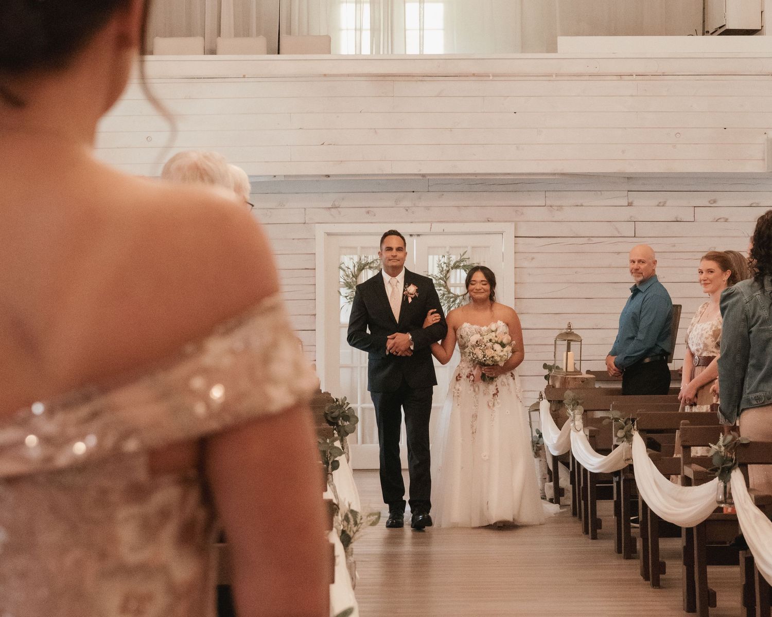 Bride in white floral gown walks down chapel aisle with escort toward waiting groom.