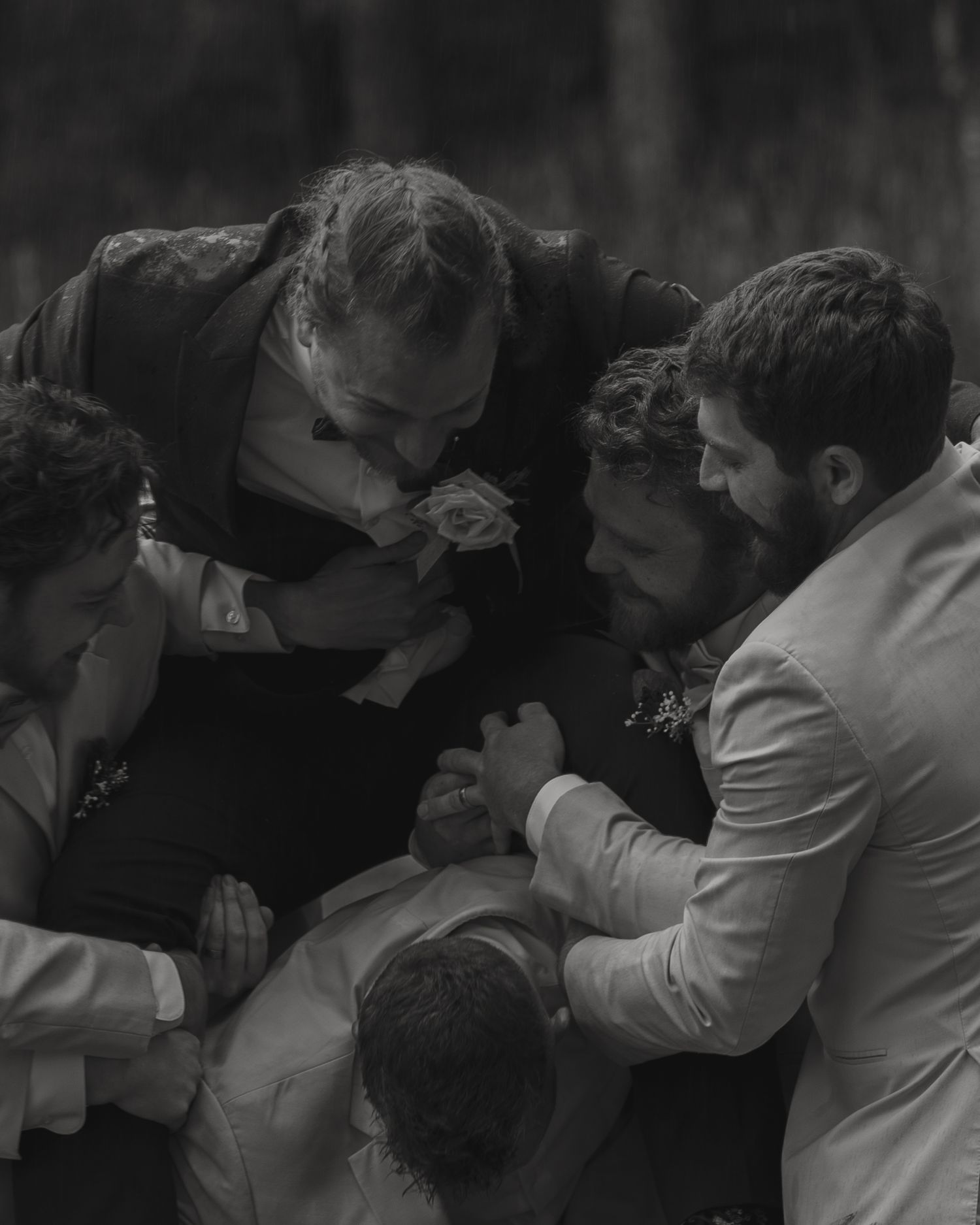 Black and white photo of groomsmen huddled together in a candid outdoor wedding moment, trying to lift him up.
