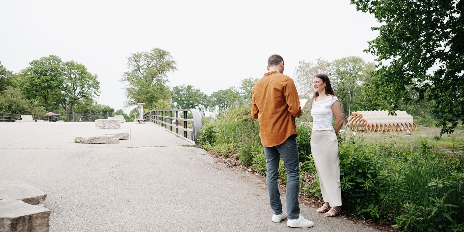 Couple standing on a scenic park path surrounded by lush green trees and gardens on a sunny day.