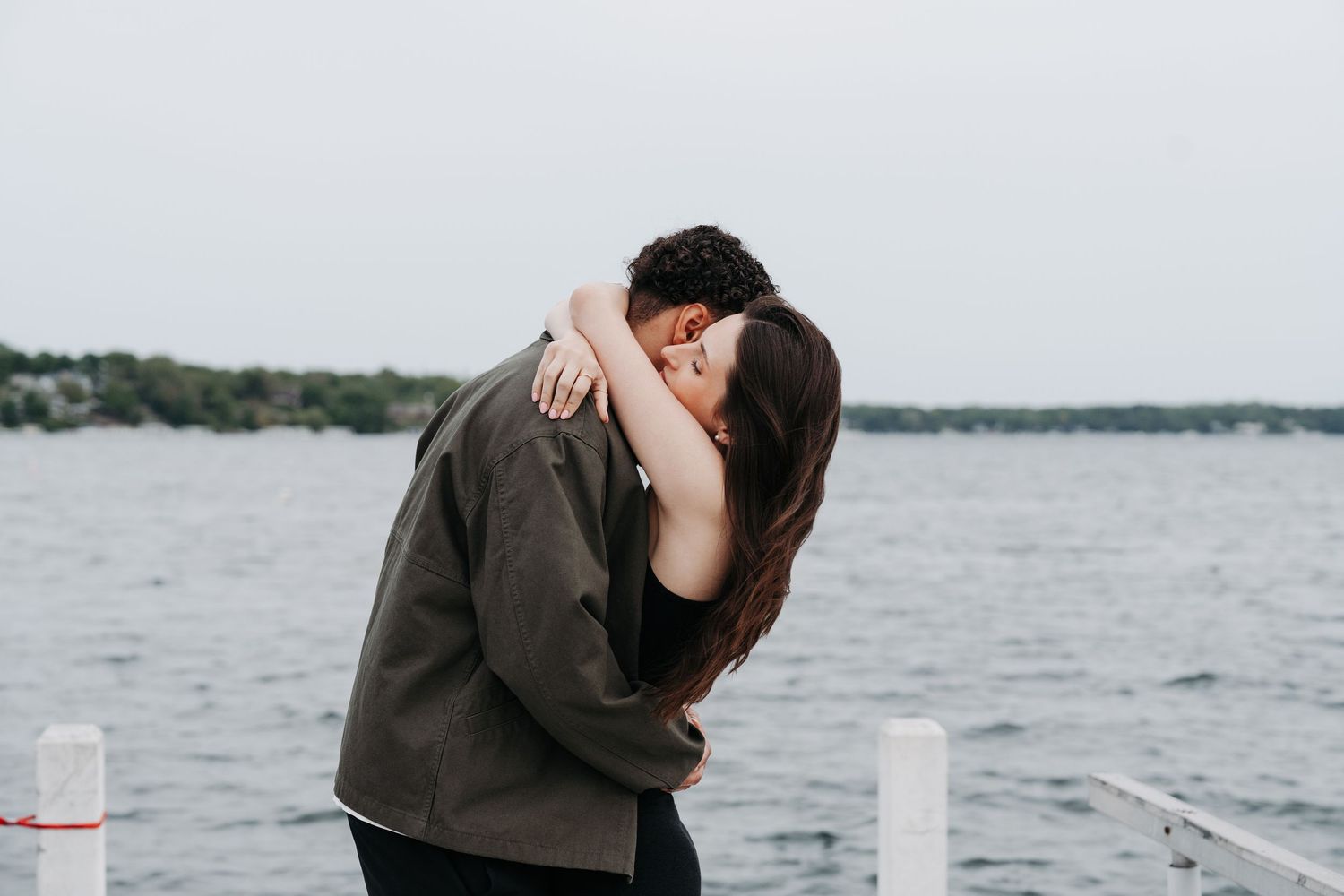 Couple sharing a romantic kiss and embrace on a waterfront dock with a serene lake in the background.