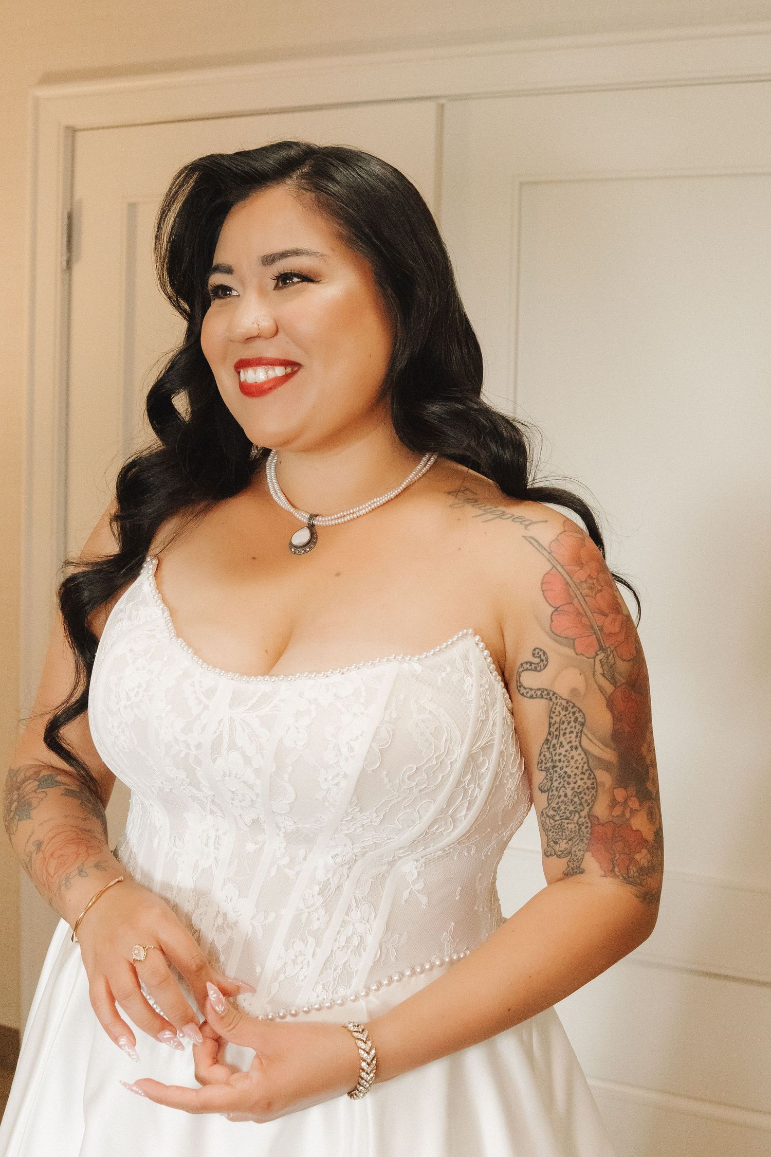 Smiling bride in white lace wedding gown with tattoos, pearl necklace, and red lips poses indoors.
