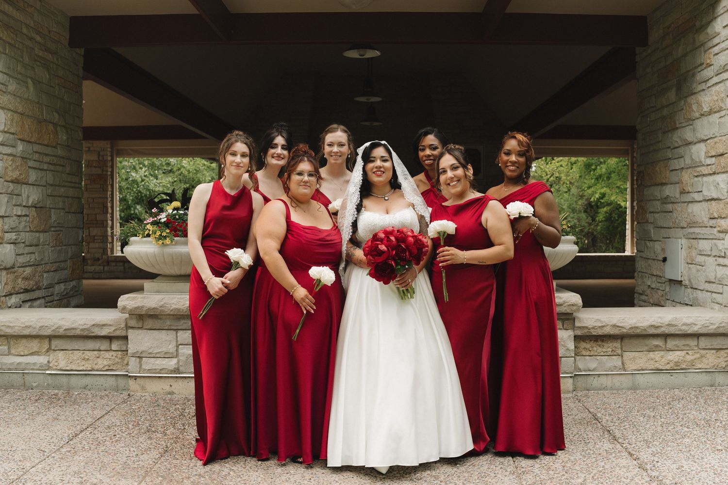 Bride in white gown poses with six bridesmaids in red dresses holding bouquets outside stone venue.