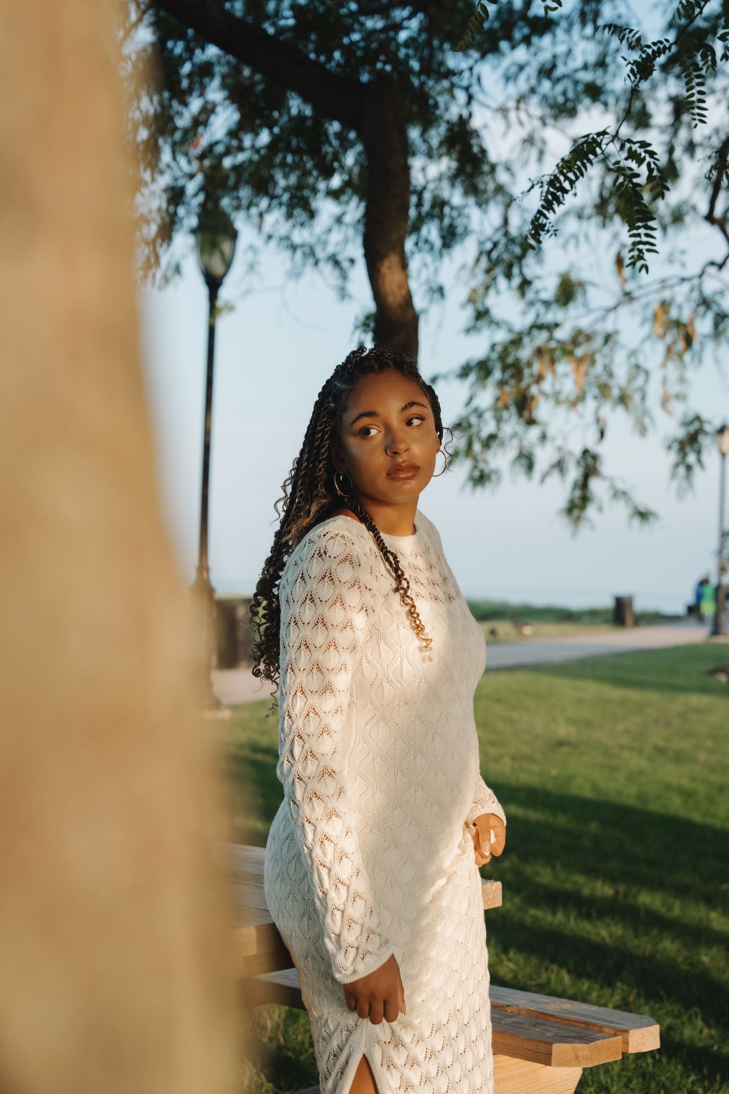 Young Black woman in white lace dress standing near park bench under tree during golden hour.