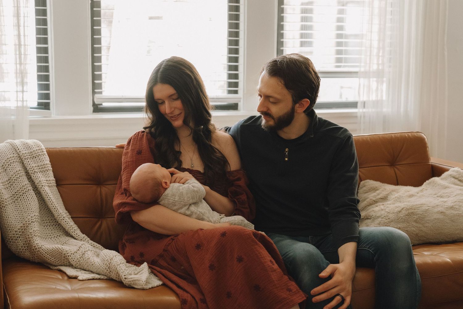 A couple sits on a brown leather sofa admiring their newborn baby wrapped in a red blanket.