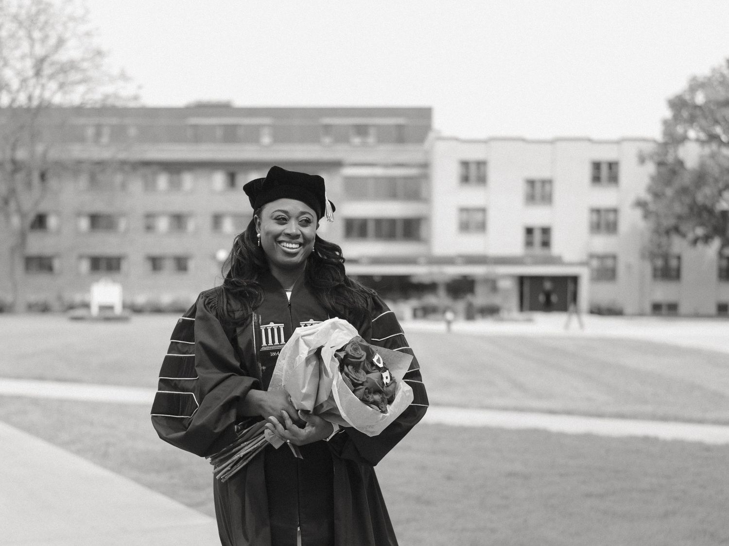 Graduate in cap and gown smiles holding flowers on campus in black and white photo.