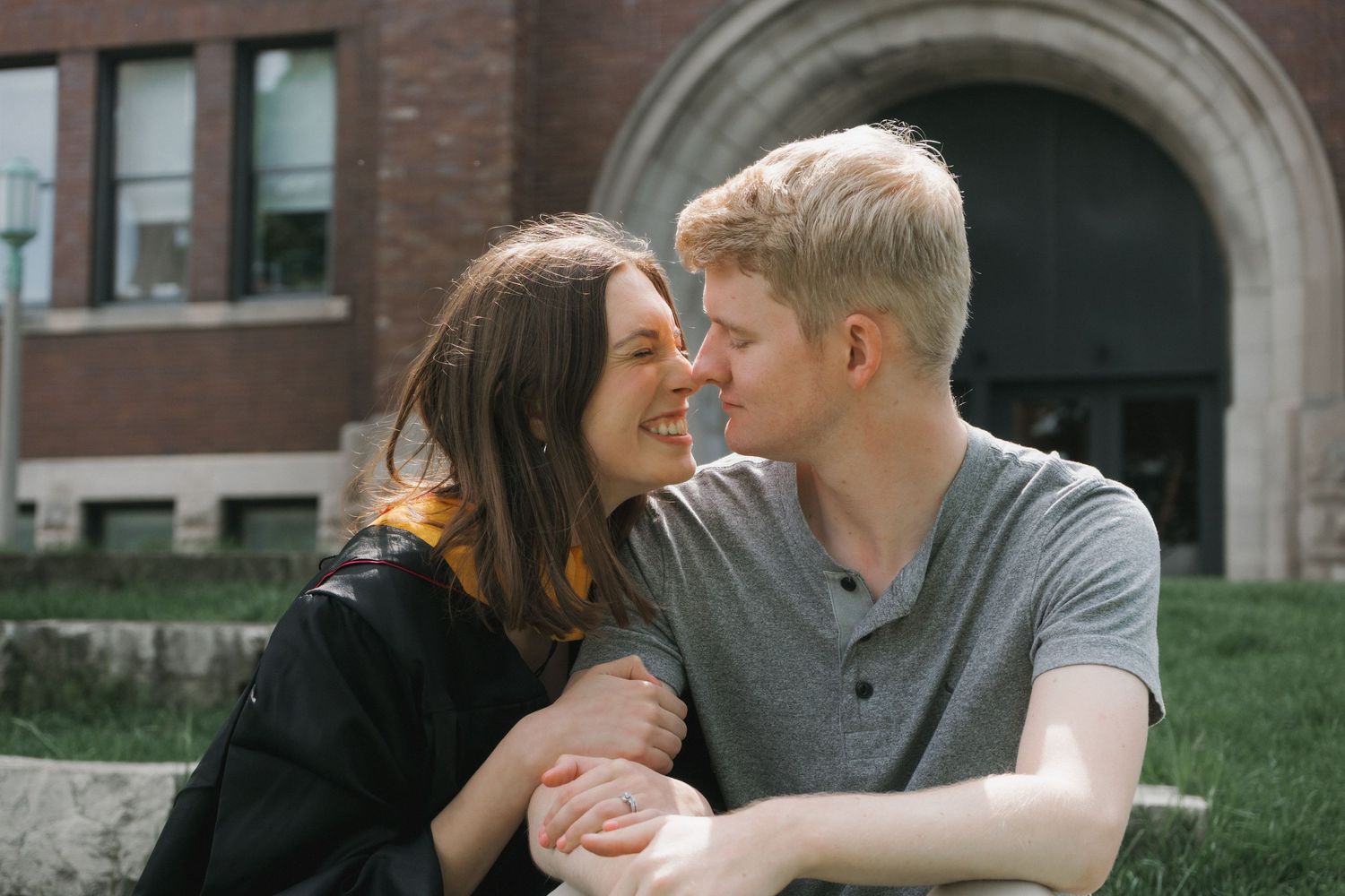 A young couple sitting on grass, smiling and gazing at each other affectionately in front of a brick building.