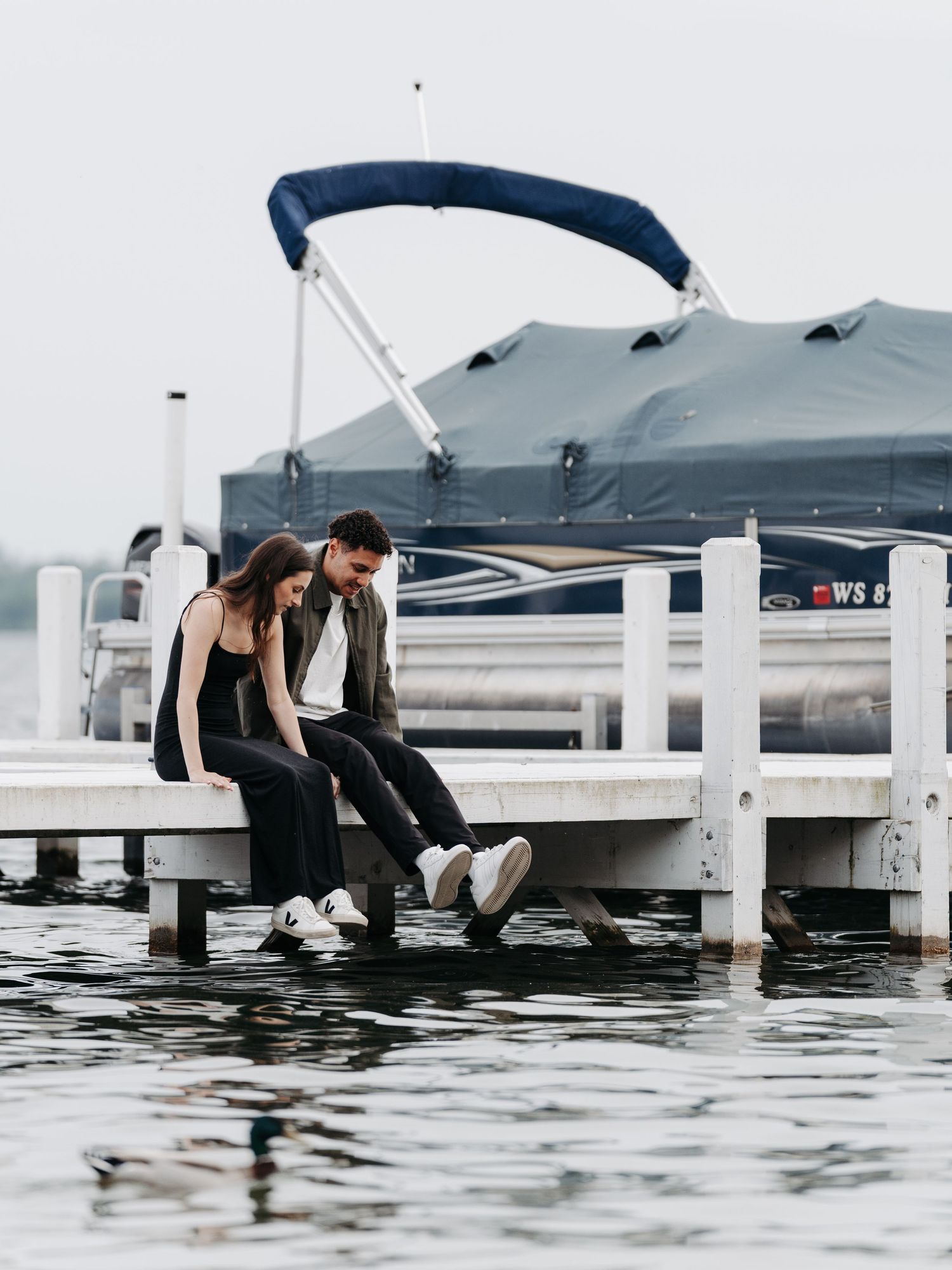 A couple sits together on a white dock beside a pontoon boat, dangling their feet over calm water on an overcast day.
