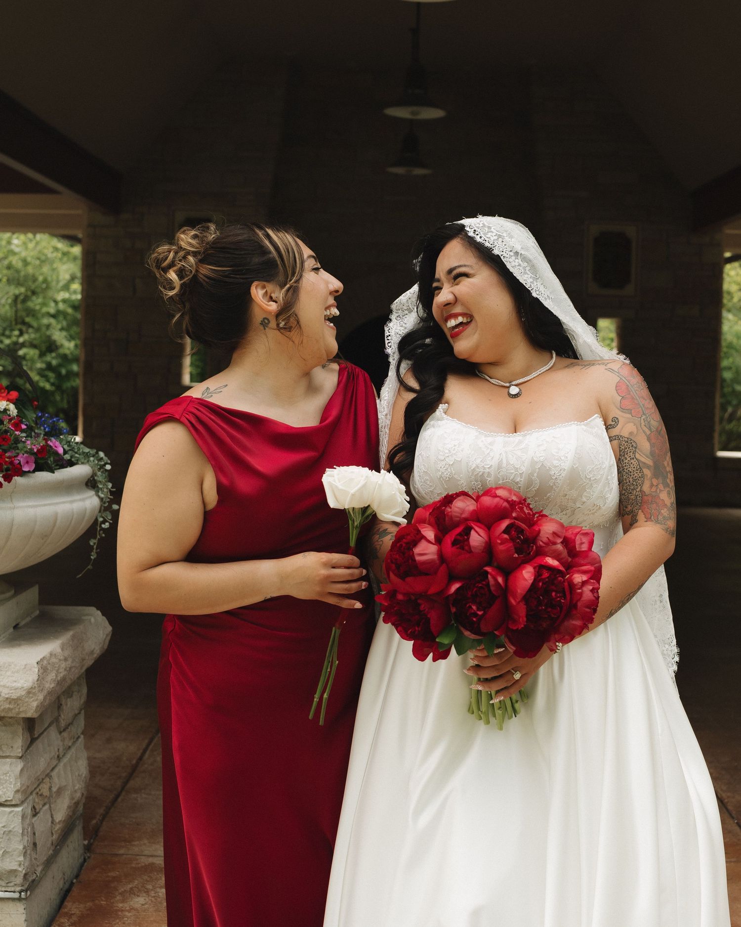Bride in white gown with red bouquet laughs with bridesmaid in red velvet dress at outdoor wedding.