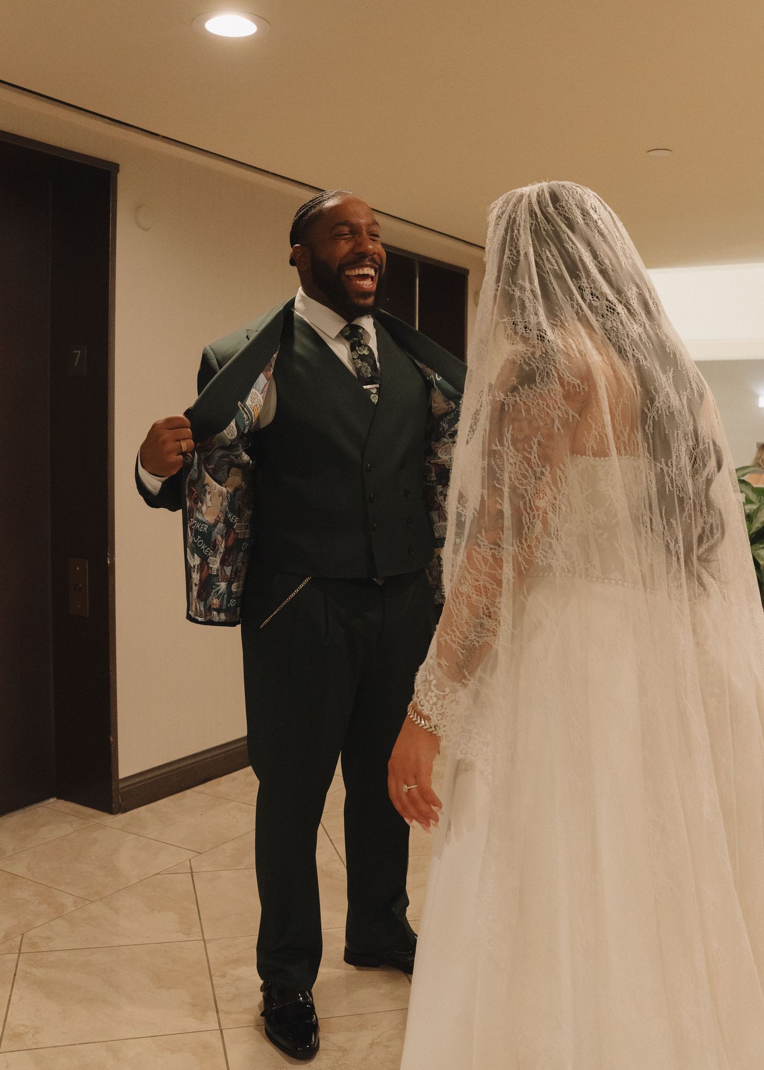Groom in dark suit laughs joyfully during first look moment with bride in white lace gown and veil.
