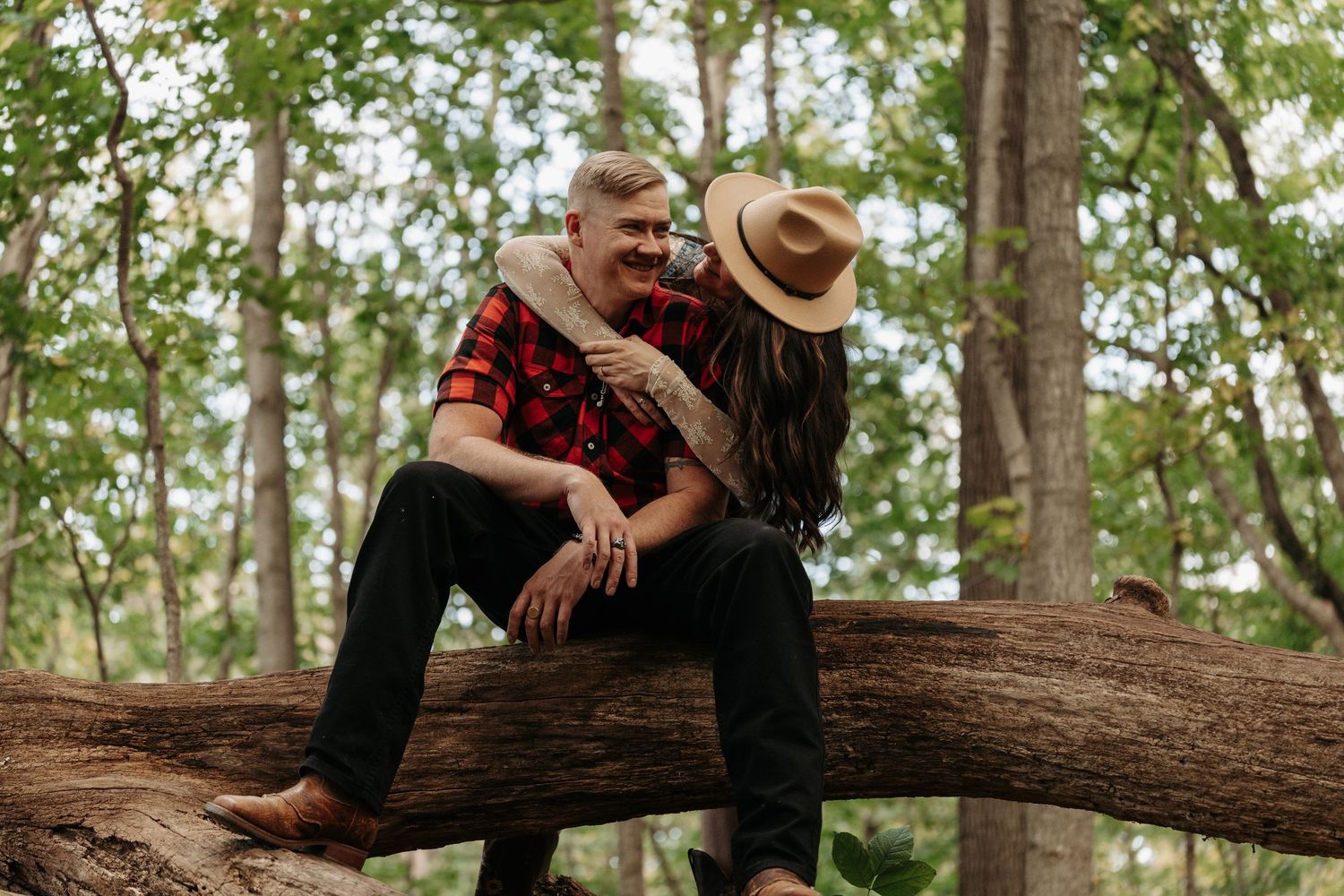 Couple sitting on a fallen log in the woods, woman nuzzling mans neck affectionately.