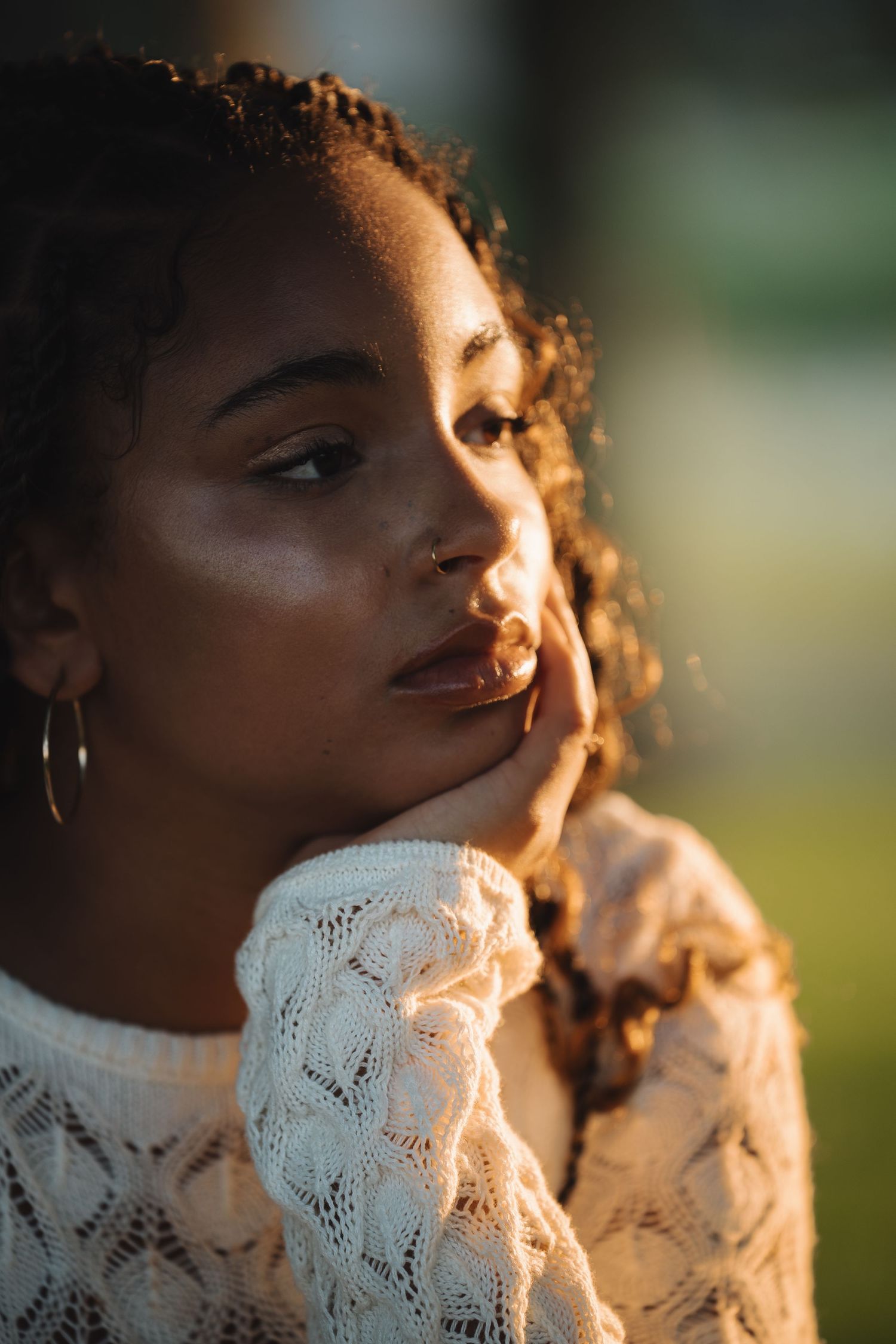 A young Black woman in a white lace top gazes thoughtfully, hand on cheek, wearing gold hoop earrings in warm sunlight.