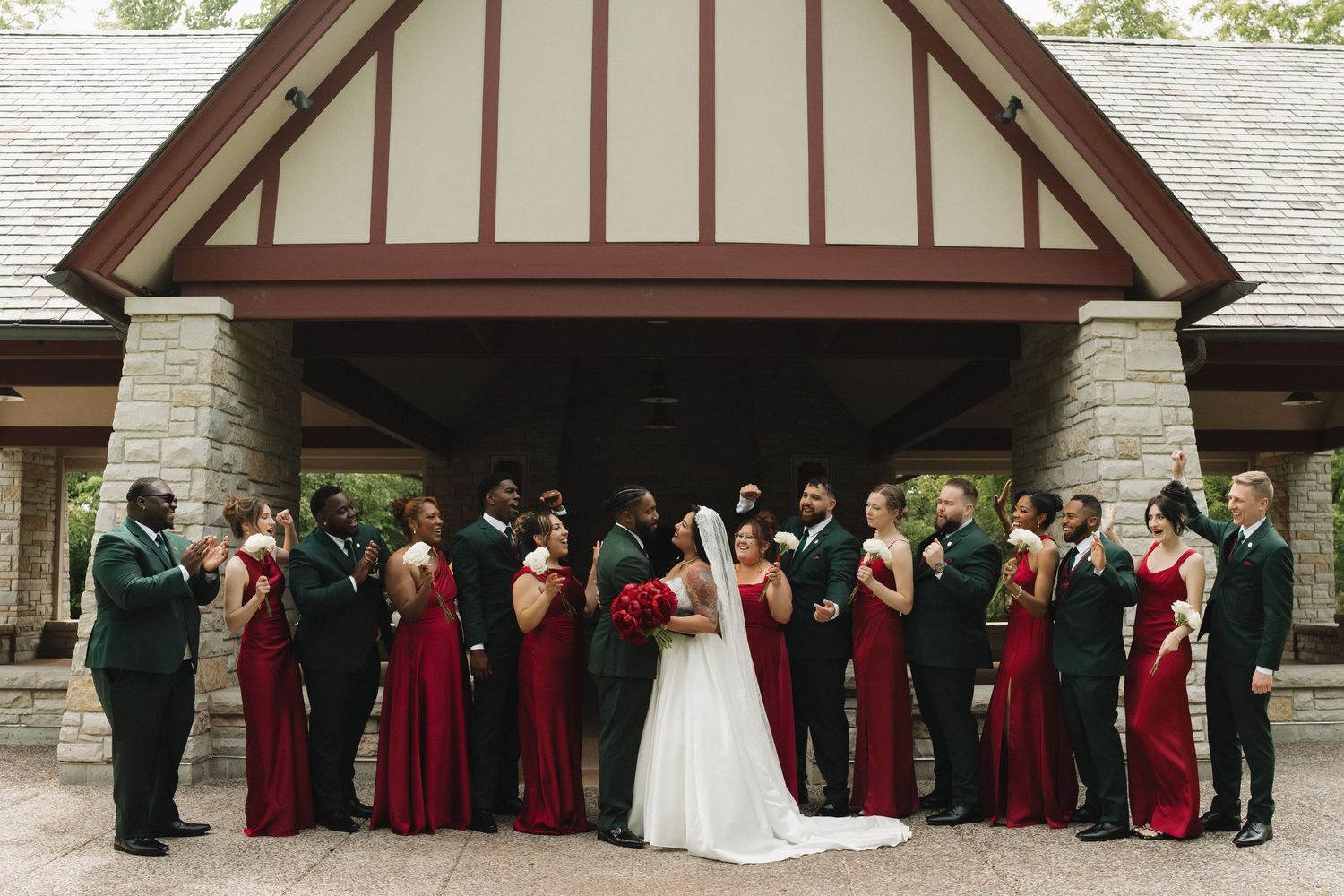 A wedding party in red and green attire poses together outside a stone venue building.
