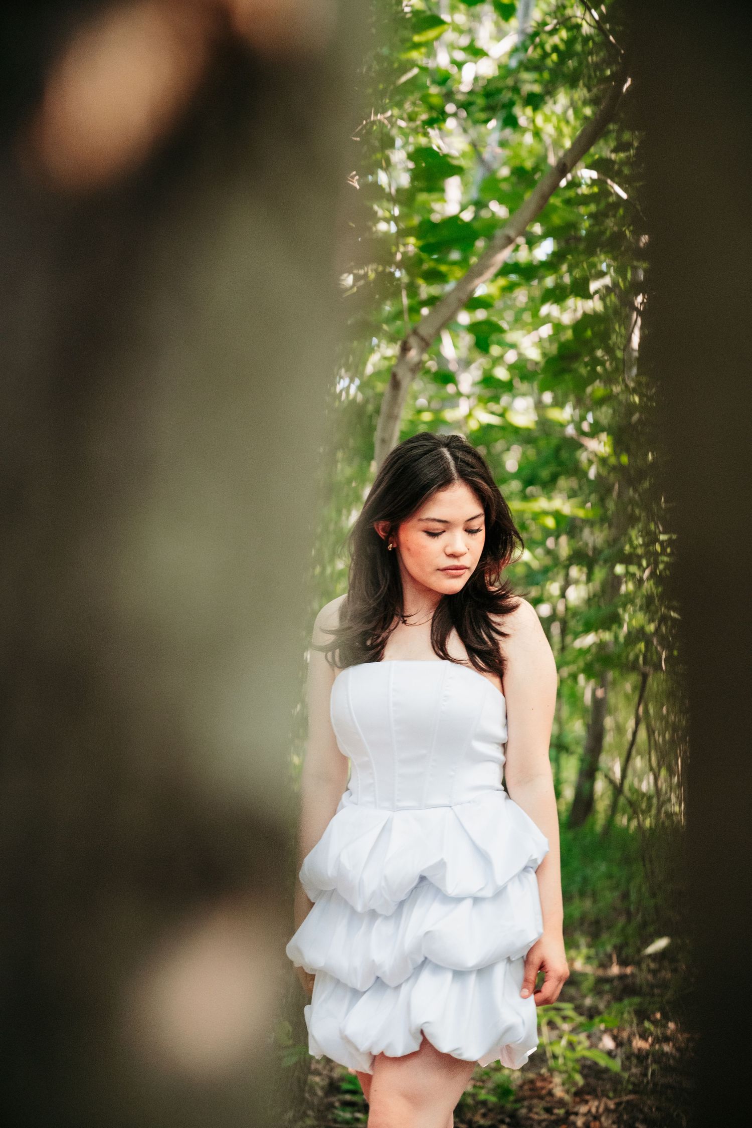 Young woman in white ruffled strapless dress standing between trees in a green forest.