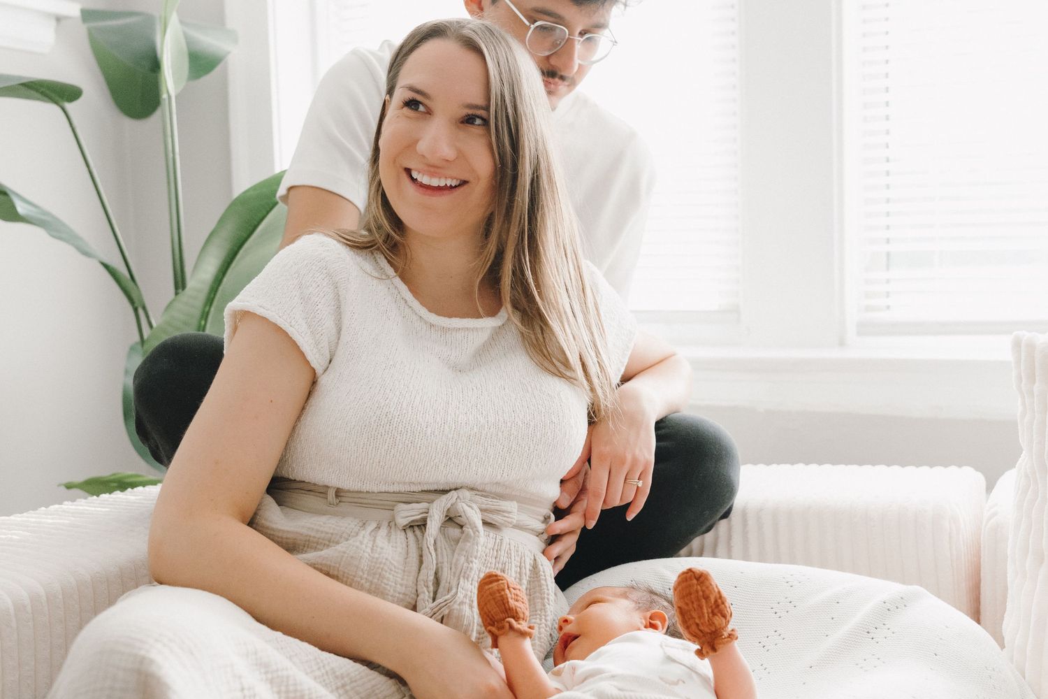 Smiling mother holding newborn baby sitting with father on white couch near green plant.