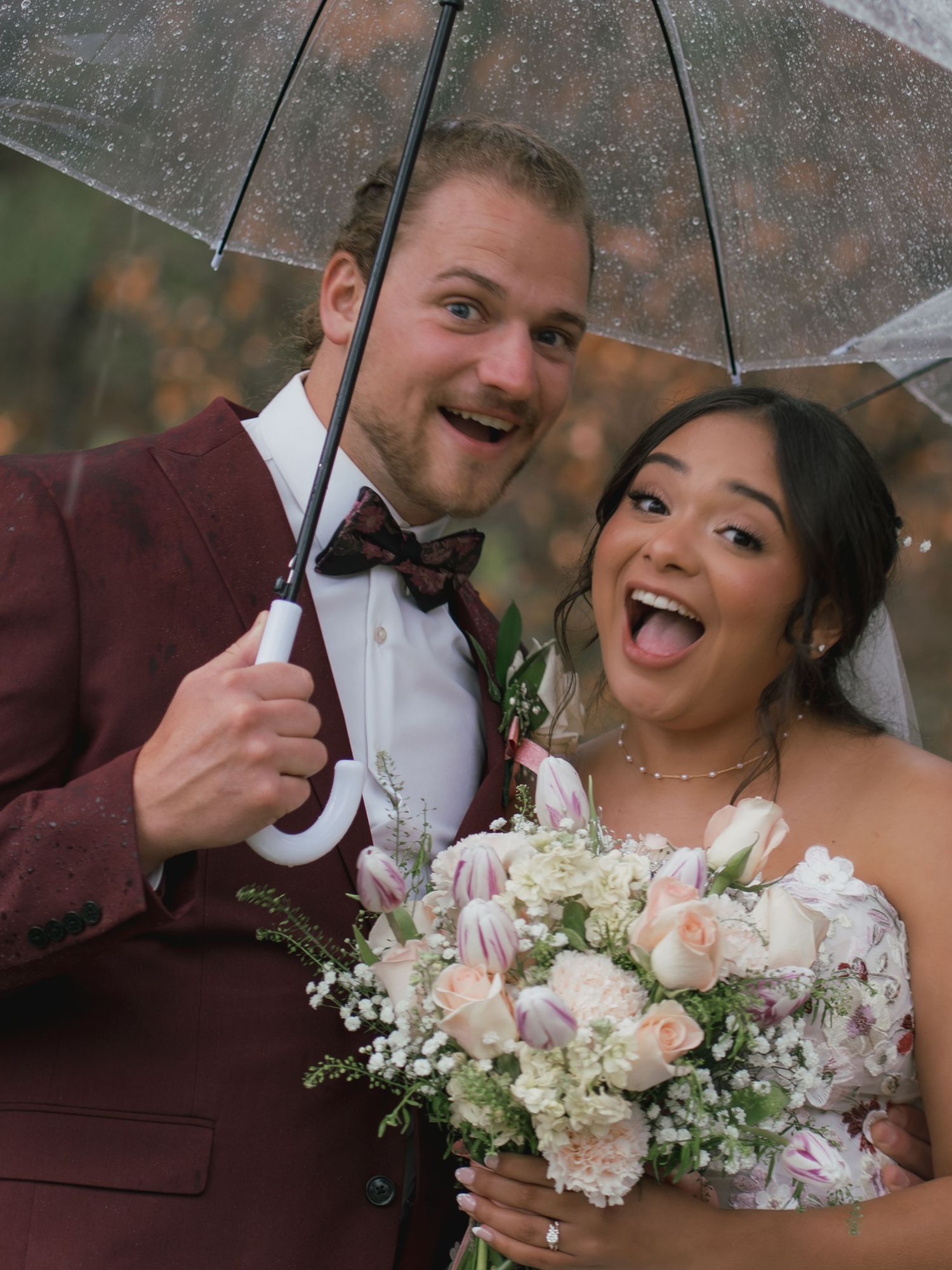 Joyful newlyweds laugh together under a clear umbrella in the rain, bride holding a tulip bouquet.