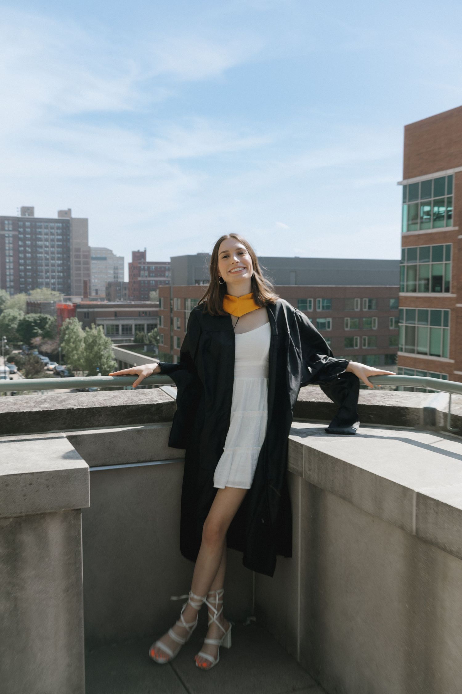 A young woman in a graduation gown and white dress poses on a rooftop terrace with city buildings in the background.