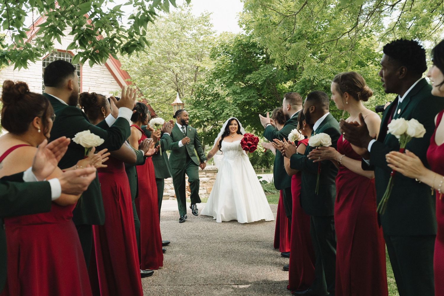 Wedding party in red dresses and dark suits applauding bride and groom walking down outdoor aisle.
