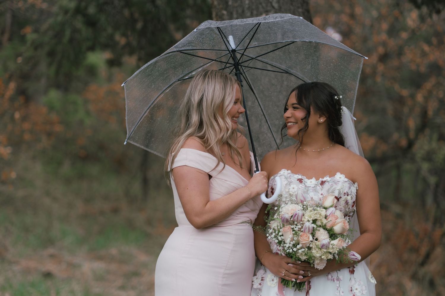 Bride and bridesmaid share a joyful moment under a clear umbrella on a rainy wedding day.