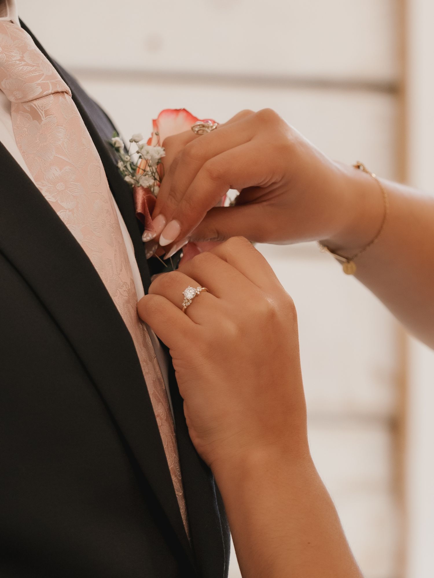 Bride with engagement ring pins pink boutonniere on father's dark suit lapel before wedding.