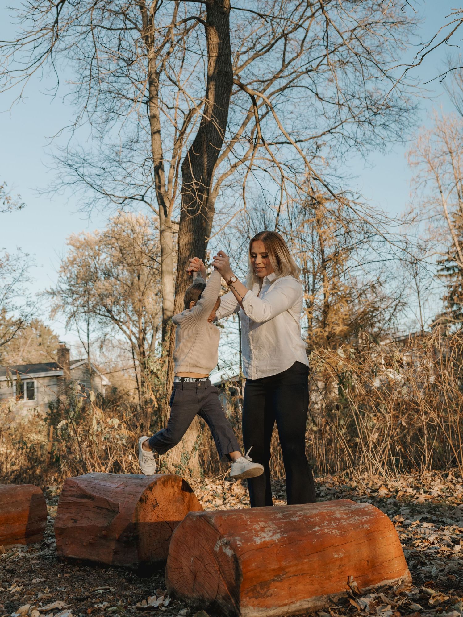 Mother helps young child balance on wooden logs in a bare autumn outdoor setting.