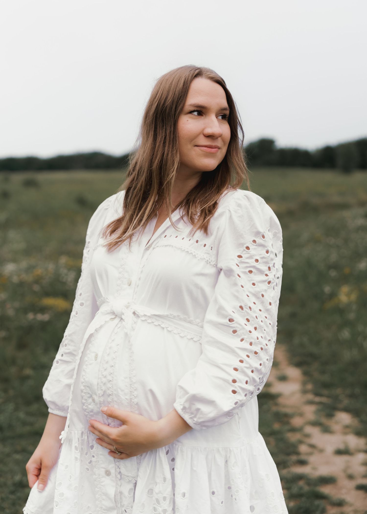 Smiling pregnant mother in white eyelet dress cradling her baby bump in a green field.