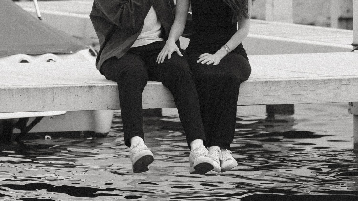 Black and white photo of a couple sitting on a dock with feet dangling over water.