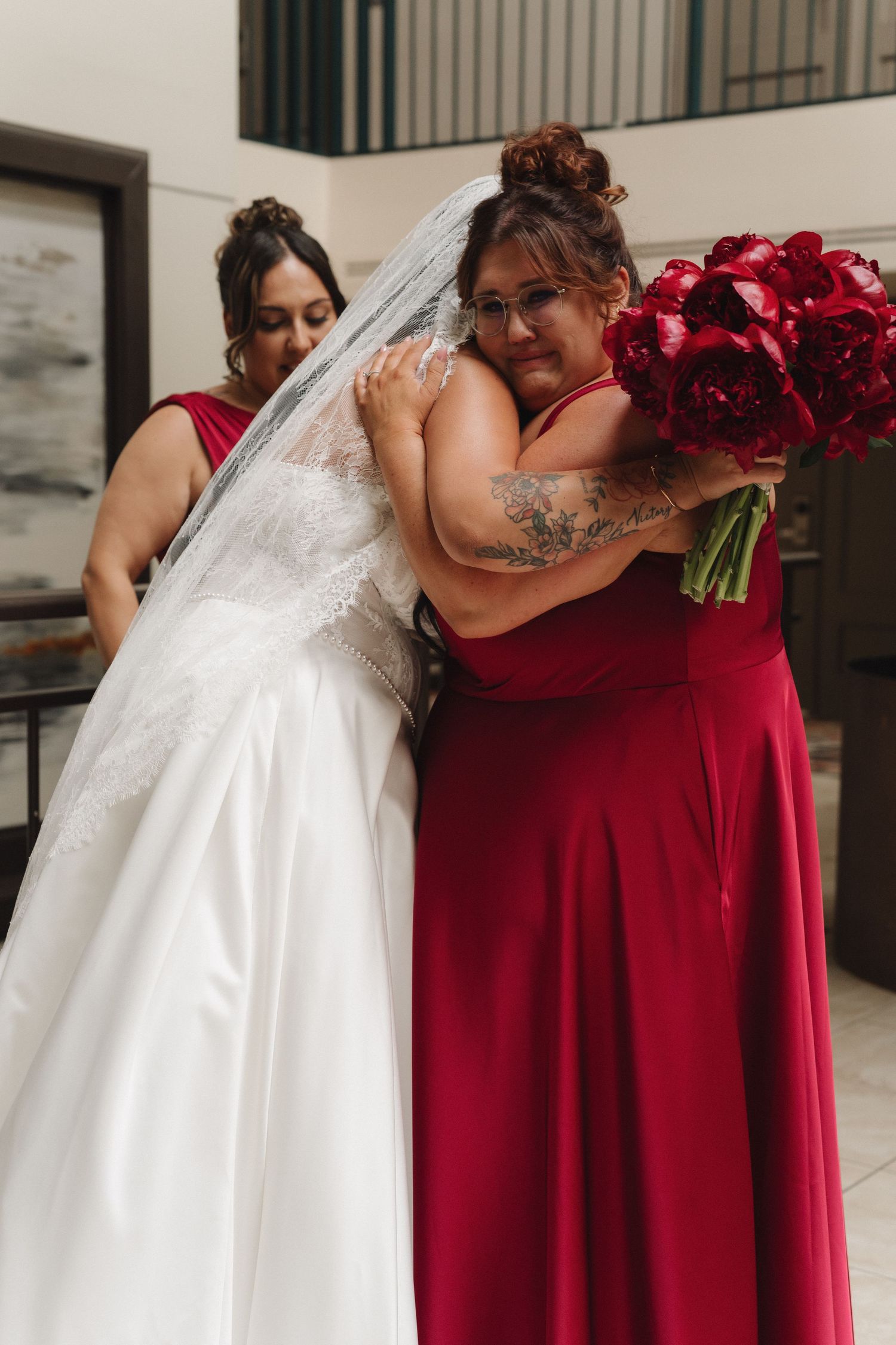 Bride in white gown and veil hugging bridesmaid in red dress holding bouquet of red peonies.