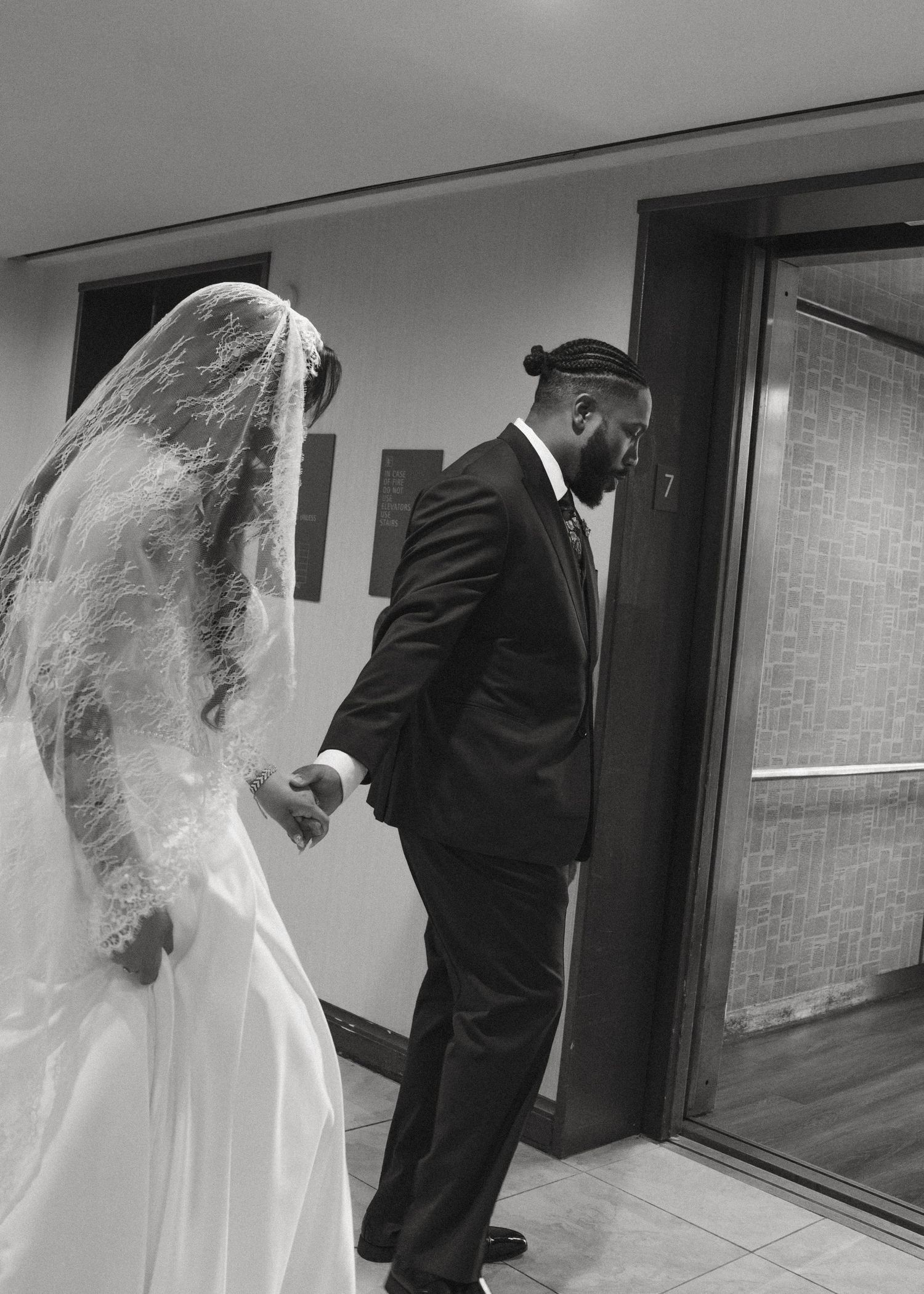 Black and white photo of a groom in a suit holding hands with a bride in a white gown and veil at their wedding.