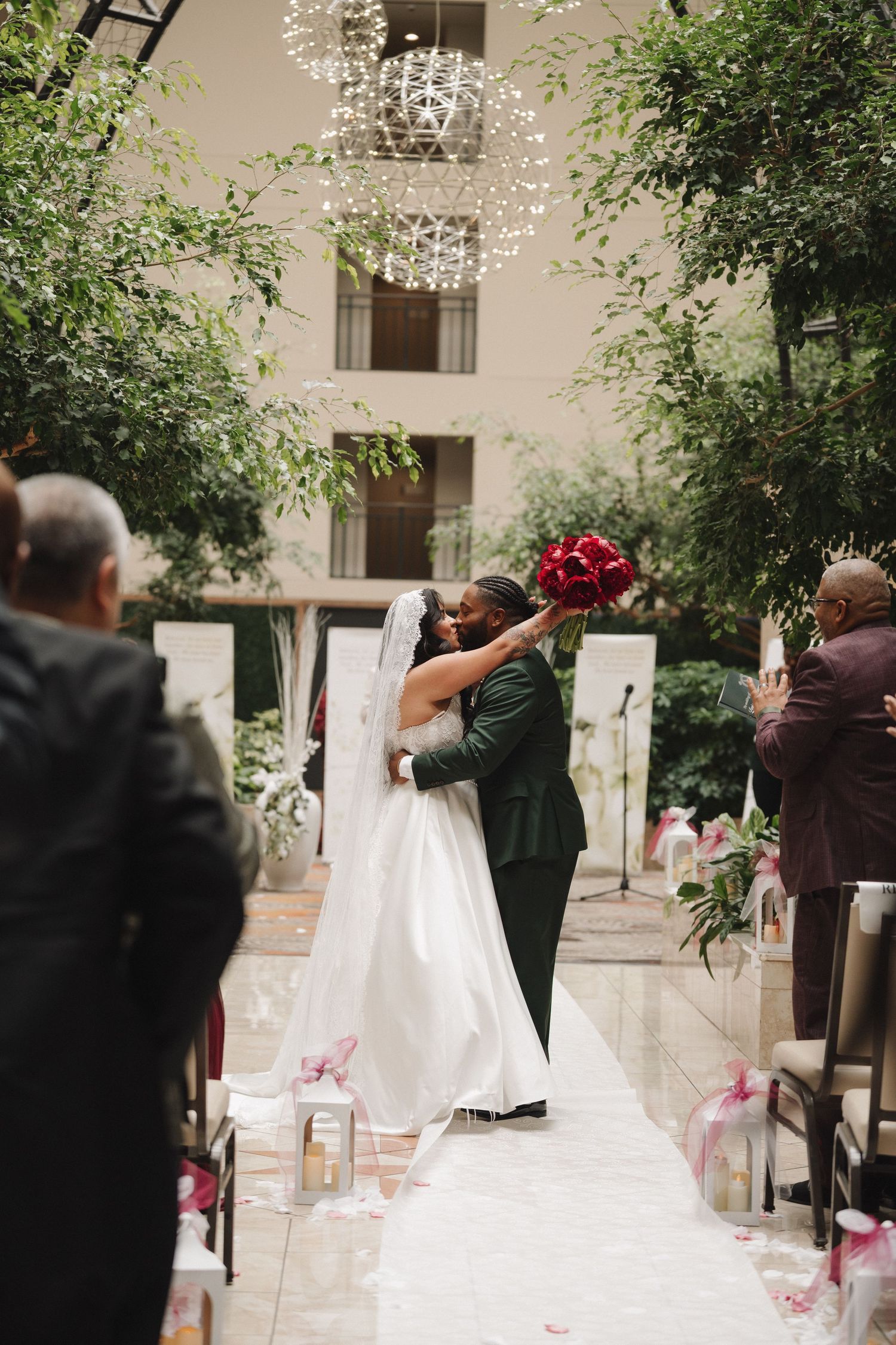 Couple shares first kiss at indoor wedding ceremony with lush greenery, chandelier, and red rose bouquet.