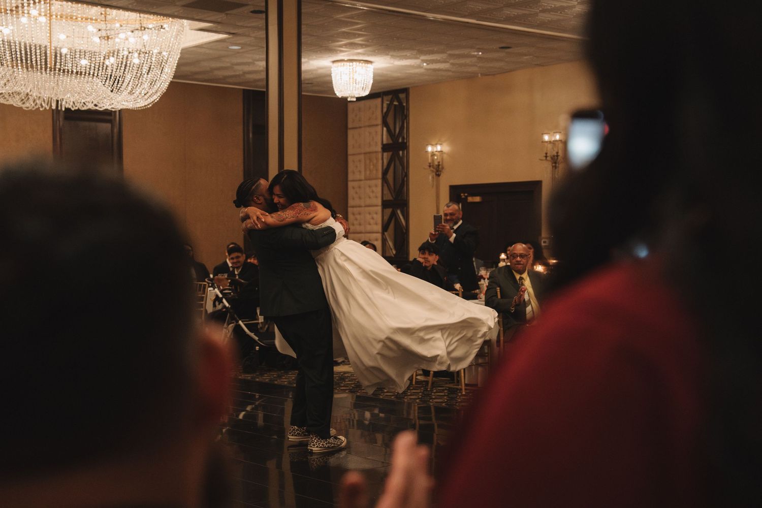 Groom lifts bride during romantic first dance in elegant ballroom with chandelier lighting at wedding reception.