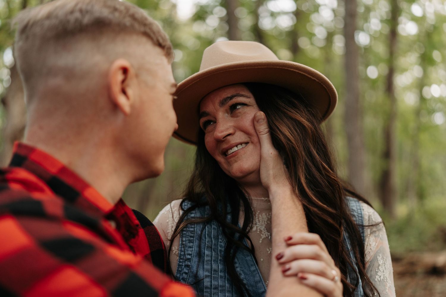 Smiling woman in a tan hat, her partner touches her face tenderly in a sunlit forest.