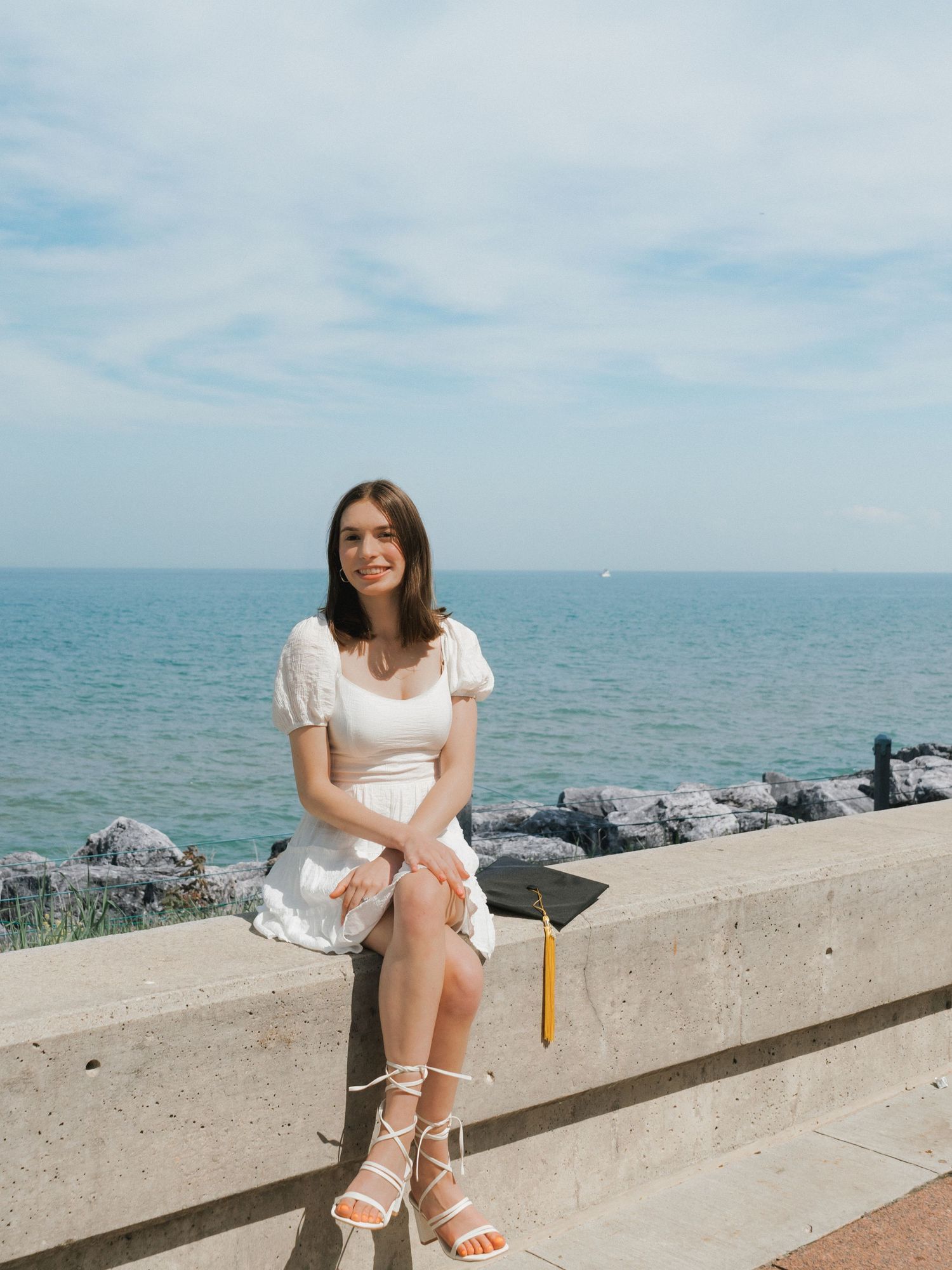A young woman in a white dress sits on a stone wall holding a graduation cap with the ocean in the background.