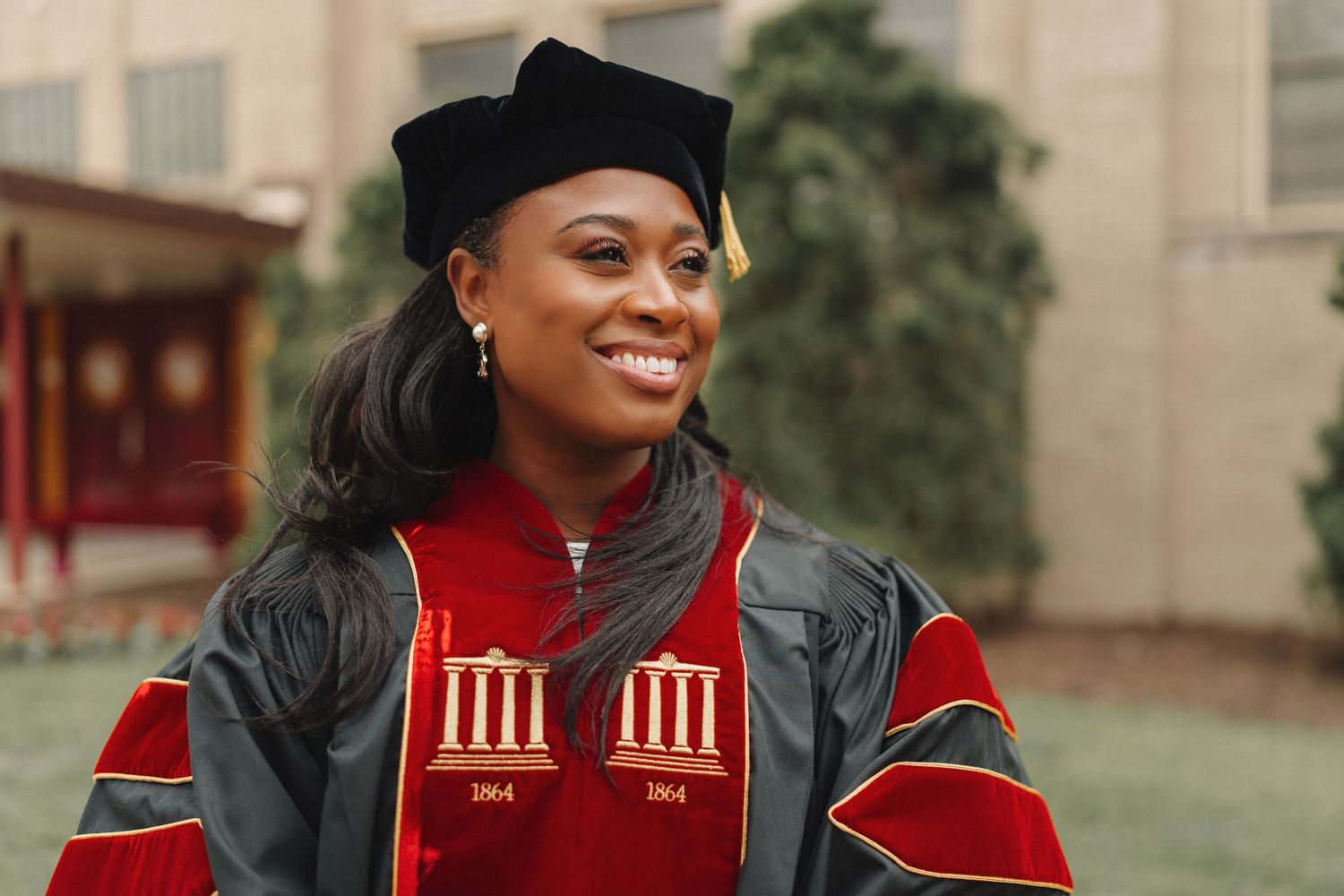 Smiling woman in red and black doctoral graduation gown and cap outdoors on campus.
