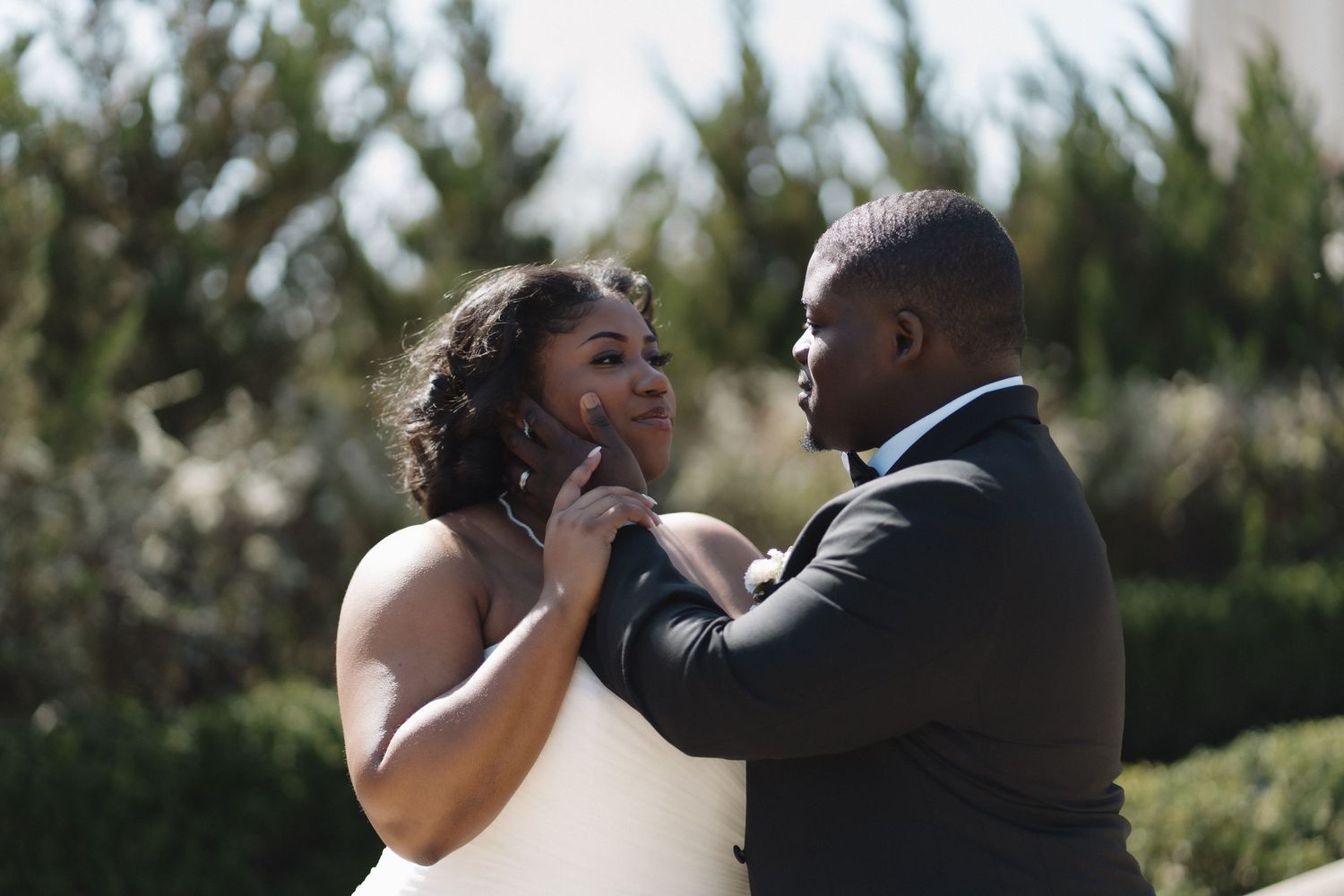 A bride and groom share a tender moment outdoors, with the groom caressing his new bride.