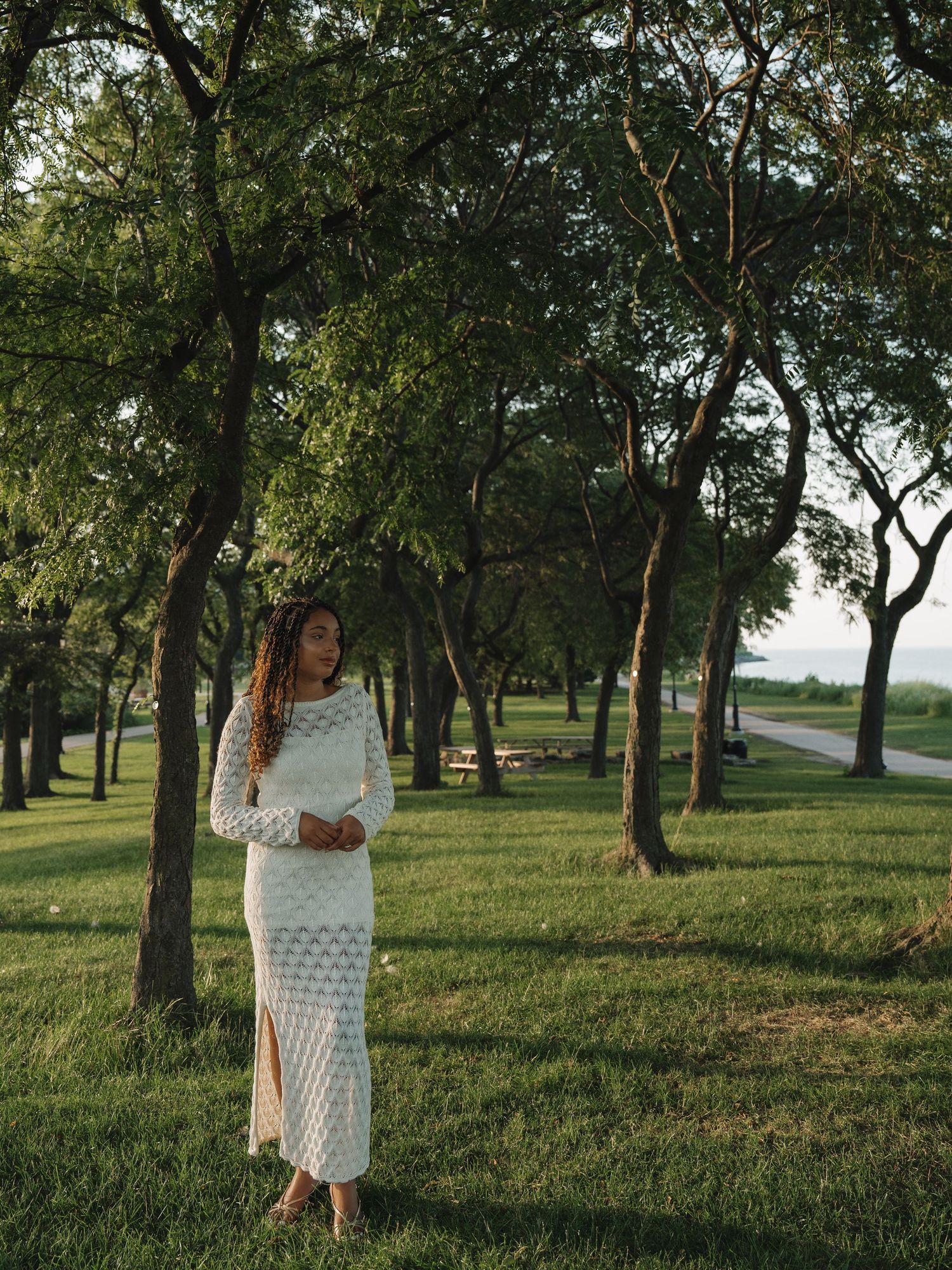 Woman in white lace maxi dress standing among green trees in a sunlit park near a waterfront.