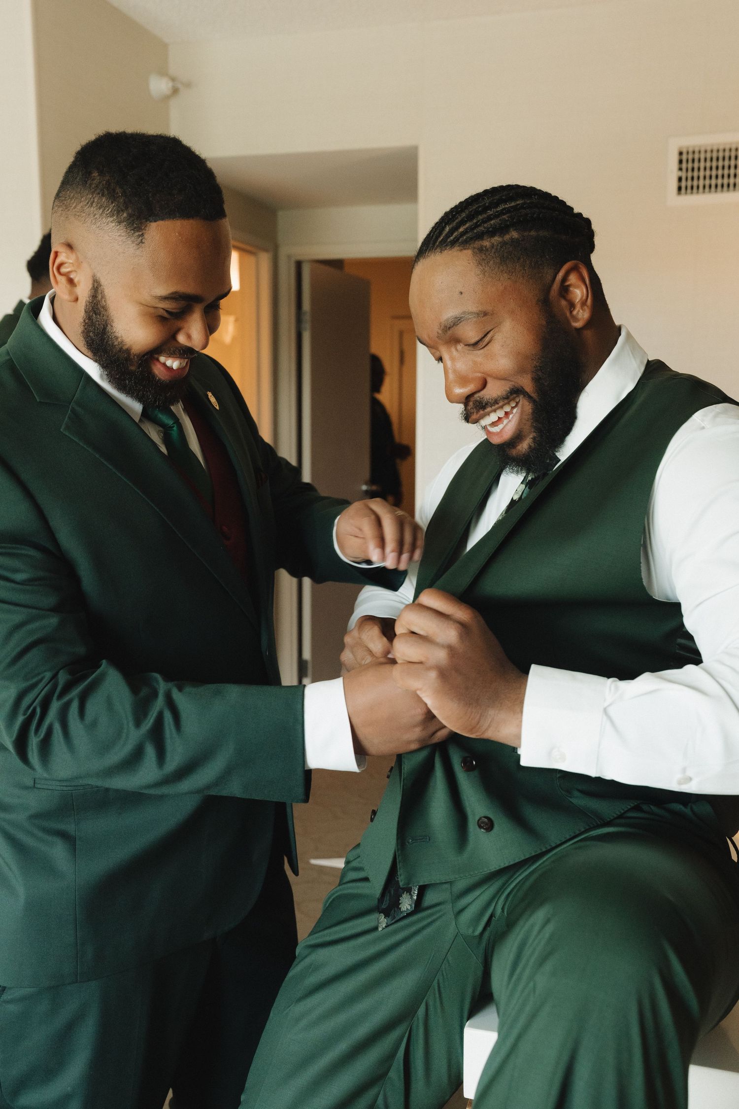 Two men in green suits smile while adjusting a vest, preparing for a wedding ceremony indoors.
