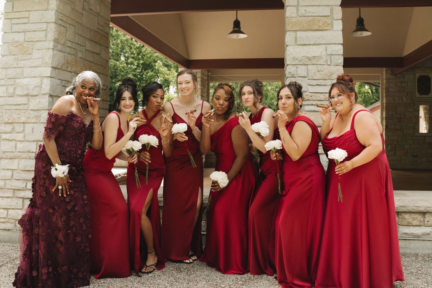 Seven bridesmaids in red gowns pose playfully holding white bouquets under a stone pavilion outdoors.