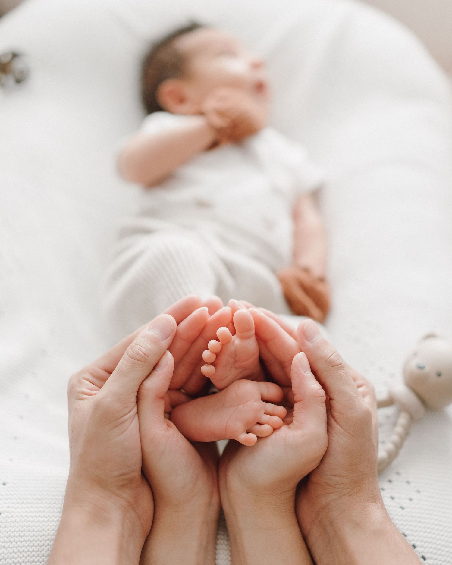 Two parents cupping newborn baby's tiny feet in their hands while baby sleeps on white surface.