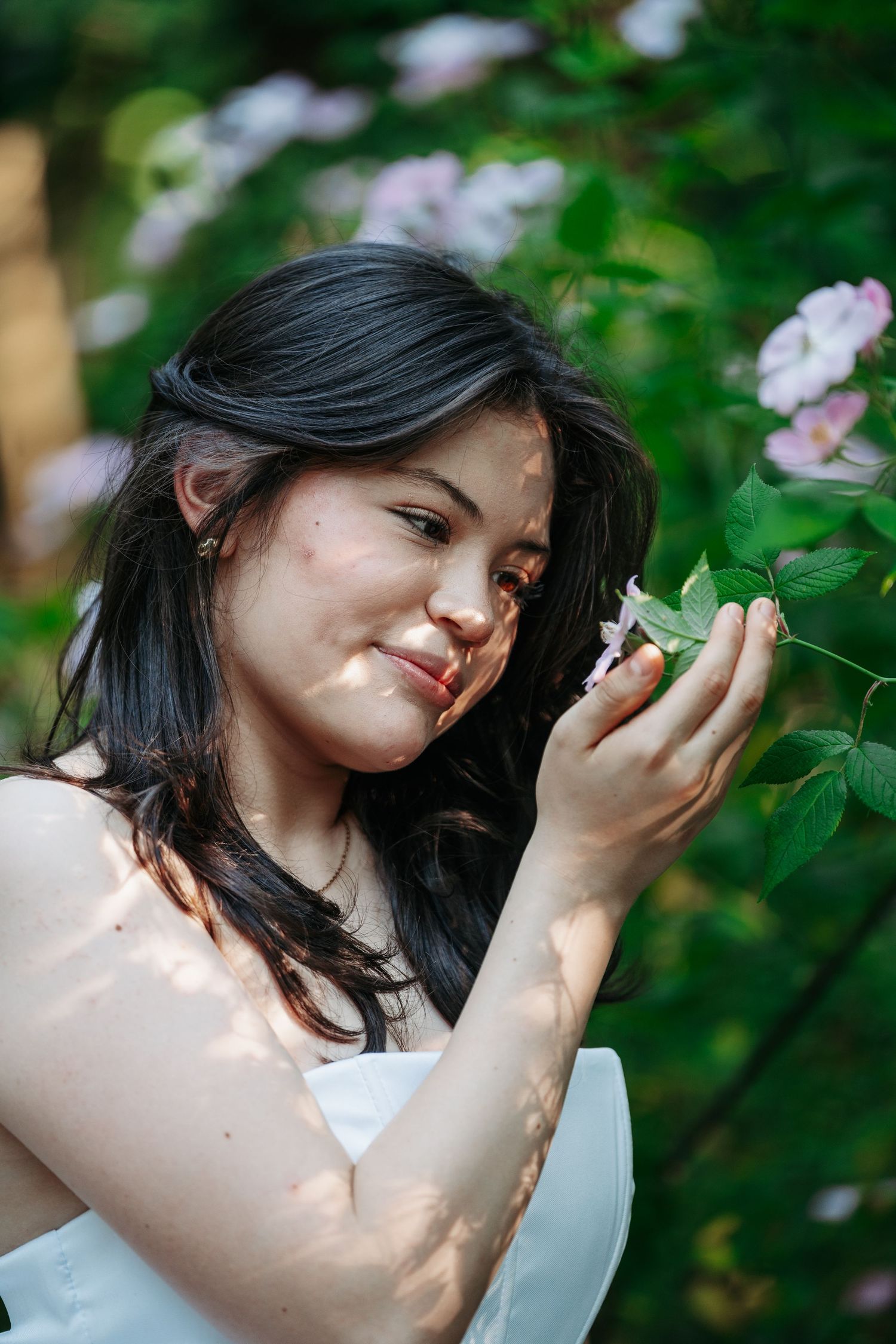 Young woman in white dress smiling while smelling pink flowers in a sunlit garden.