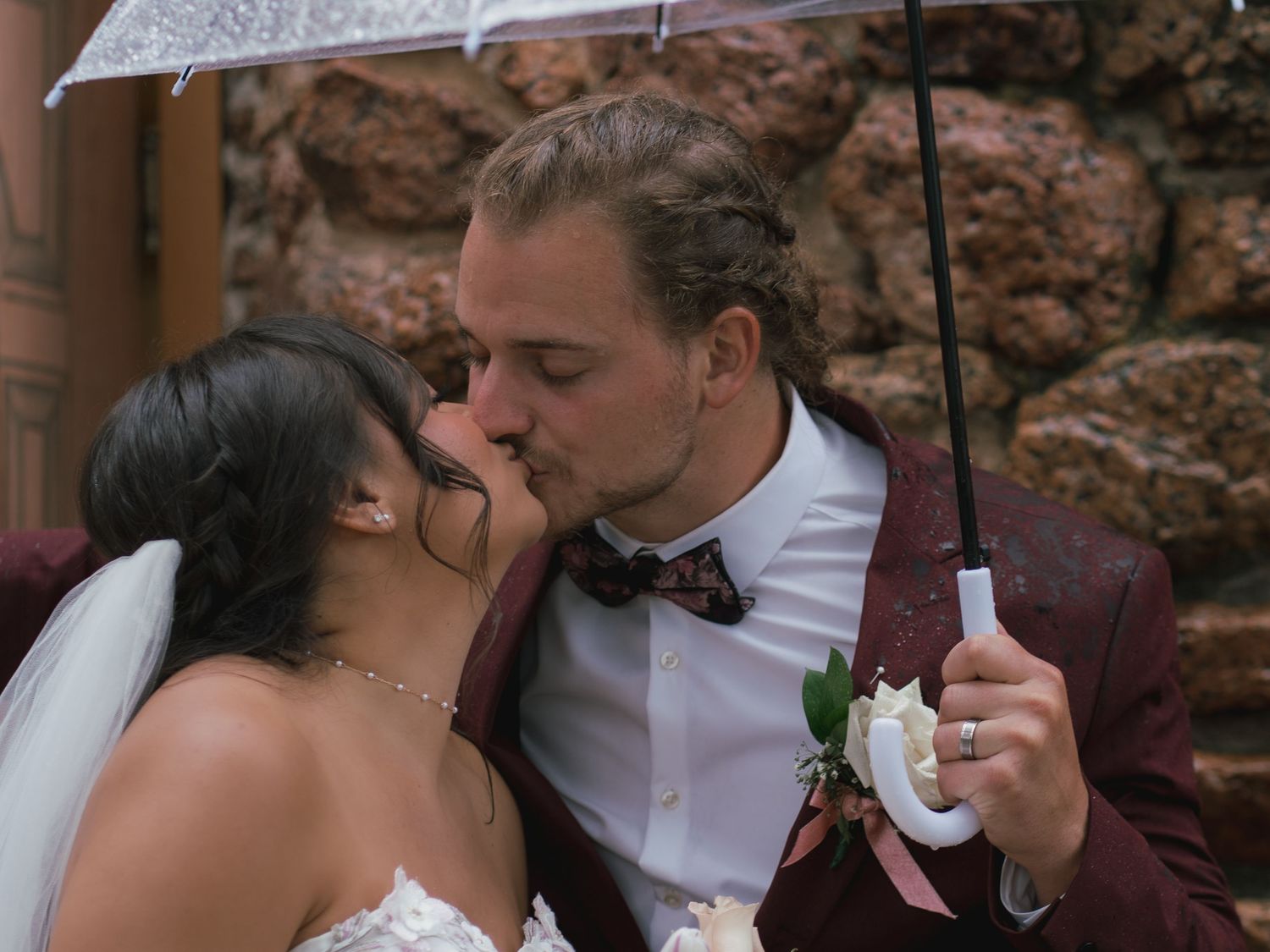 Bride and groom share a romantic kiss under a clear umbrella in the rain on their wedding day.
