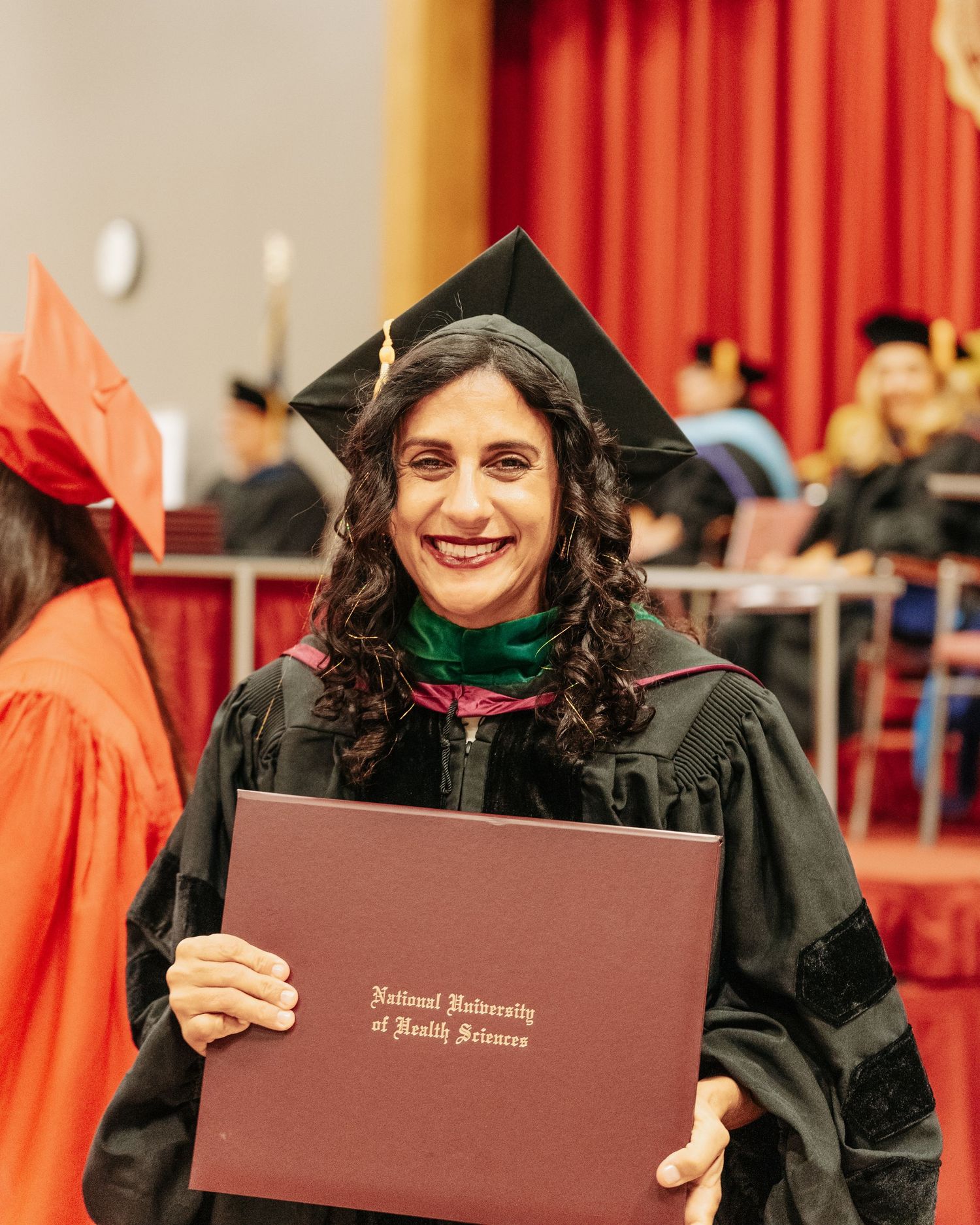 Graduate smiling in black cap and gown holding National University of Health Sciences diploma at graduation ceremony.