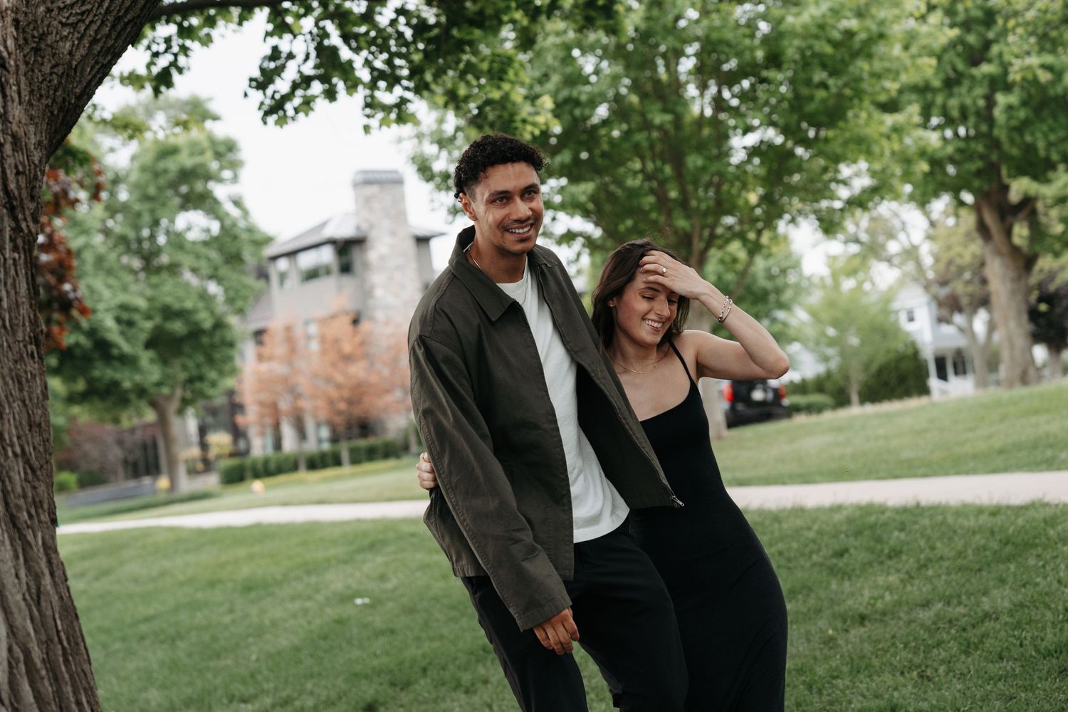 A smiling young couple stands together near a tree in a green park, the woman laughing.