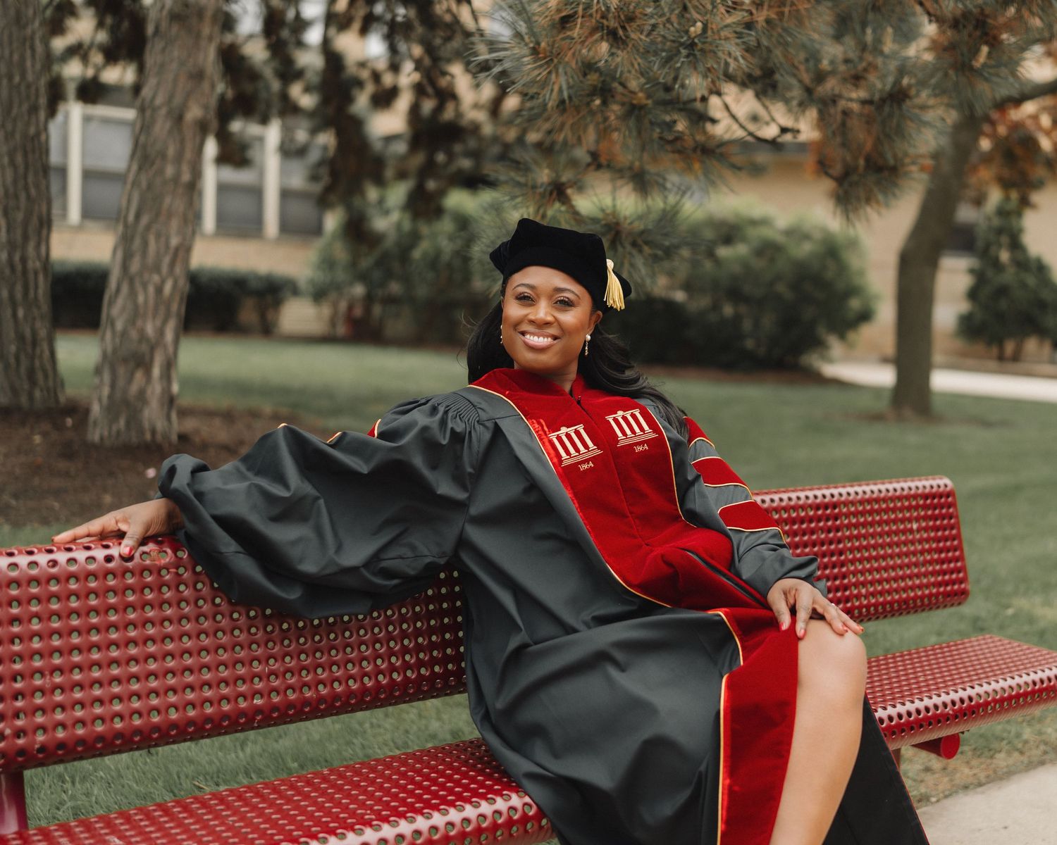 Smiling graduate in red and black doctoral regalia sitting on a red bench outdoors on a college campus.