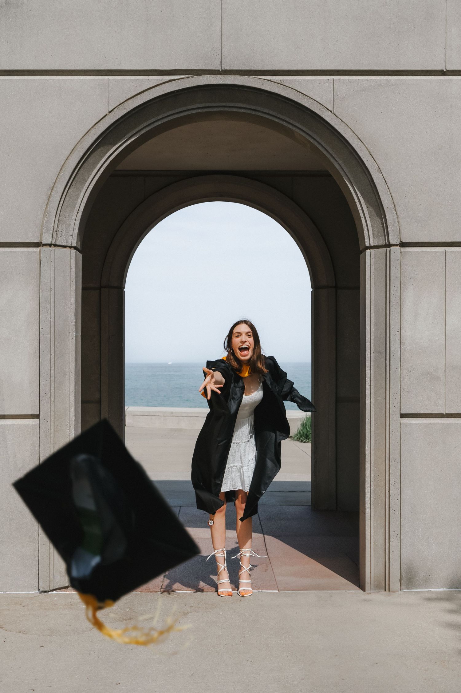 A joyful graduate in black gown tosses her cap under an arched doorway with ocean views behind her.