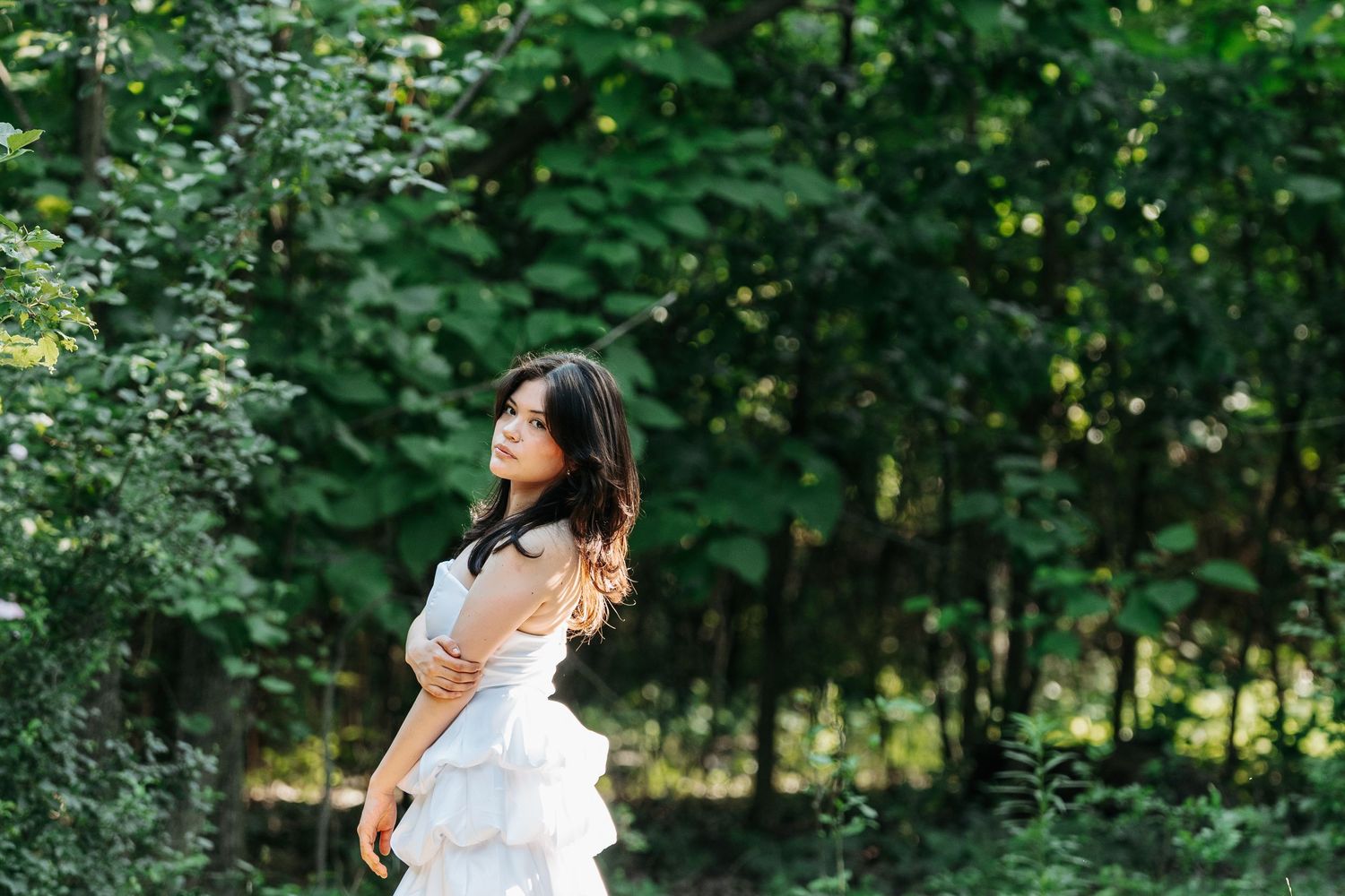 Young woman in white tiered dress twirling in lush green forest setting.