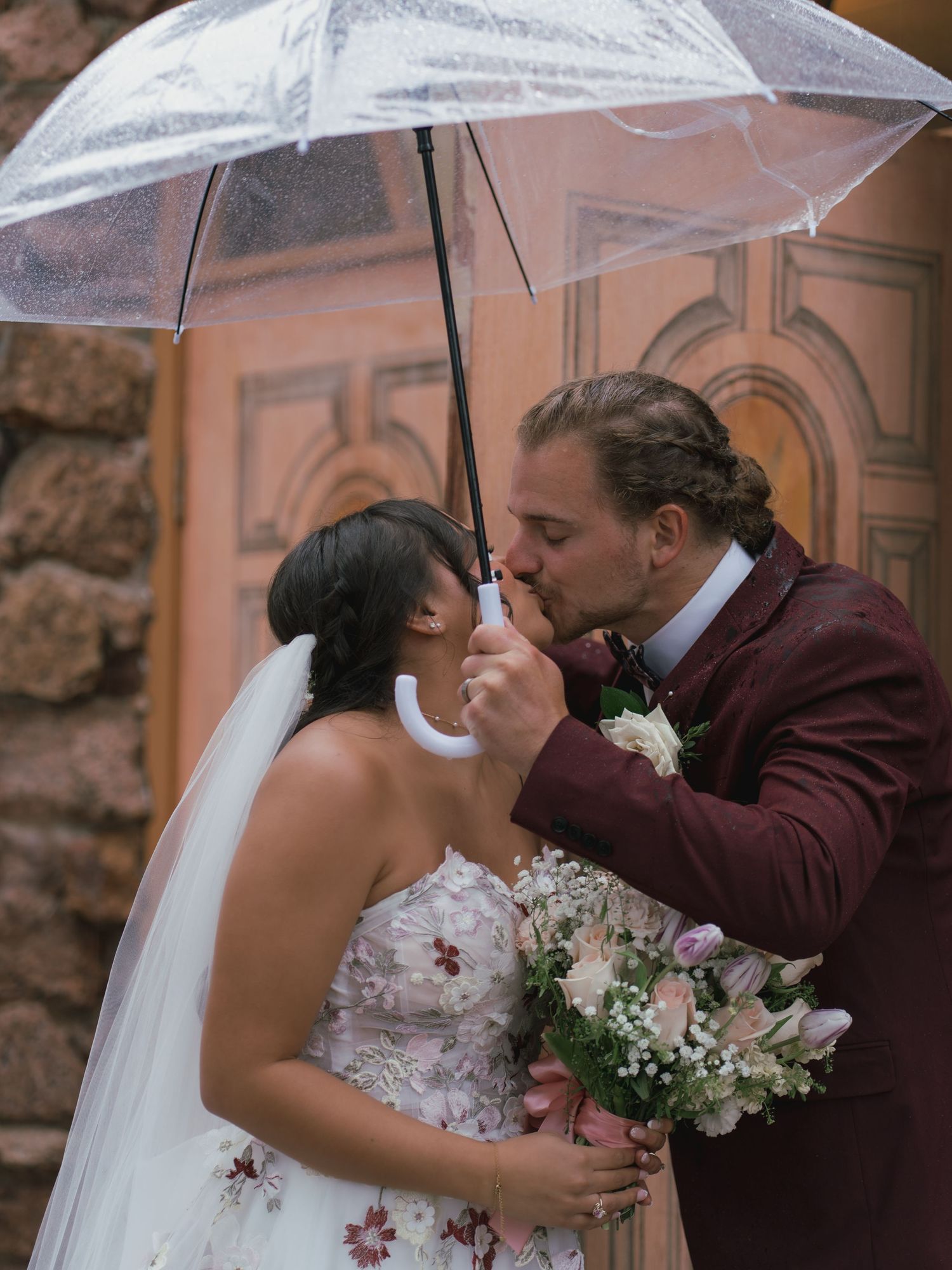 Bride and groom share a romantic kiss under a clear umbrella in the rain on their wedding day.